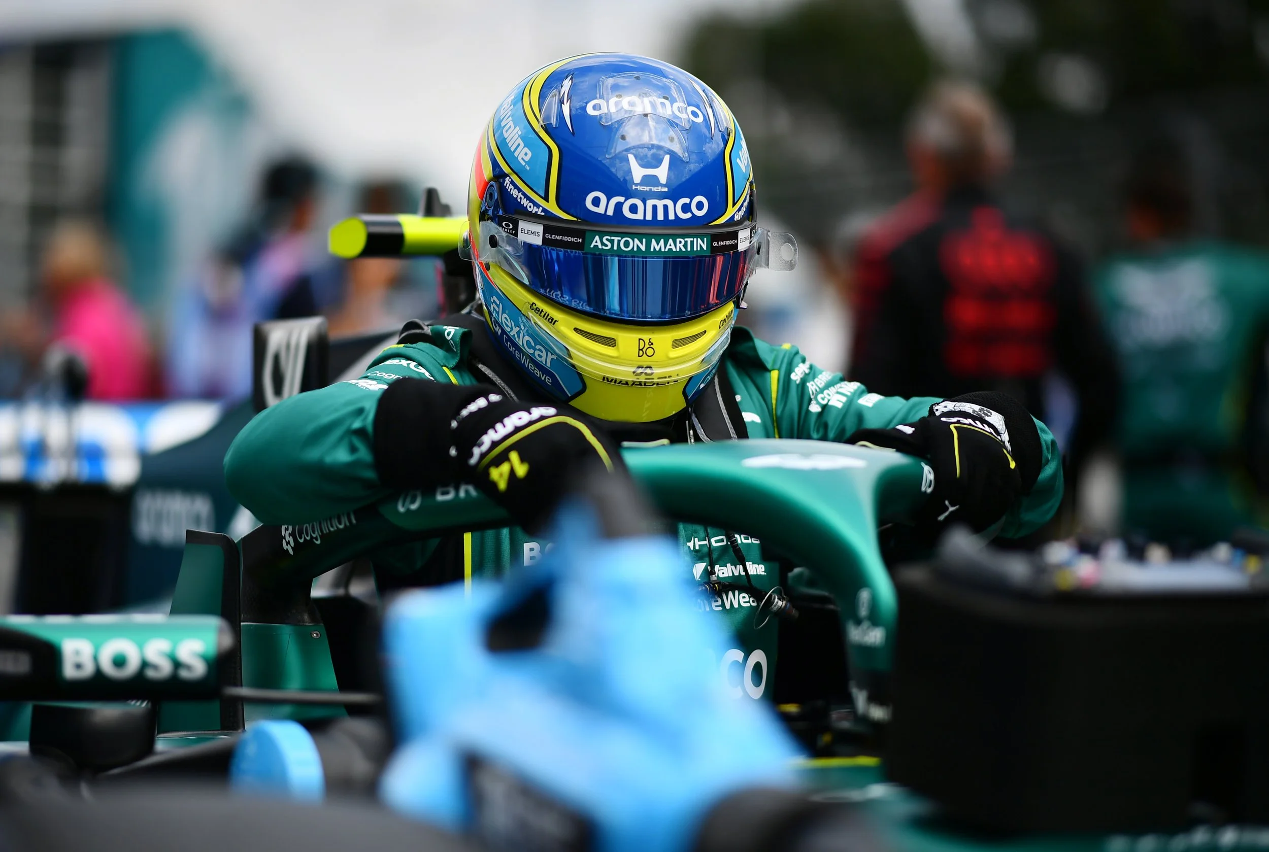 Race car driver wearing a helmet and racing suit, sitting in a green race car at a motorsport event.