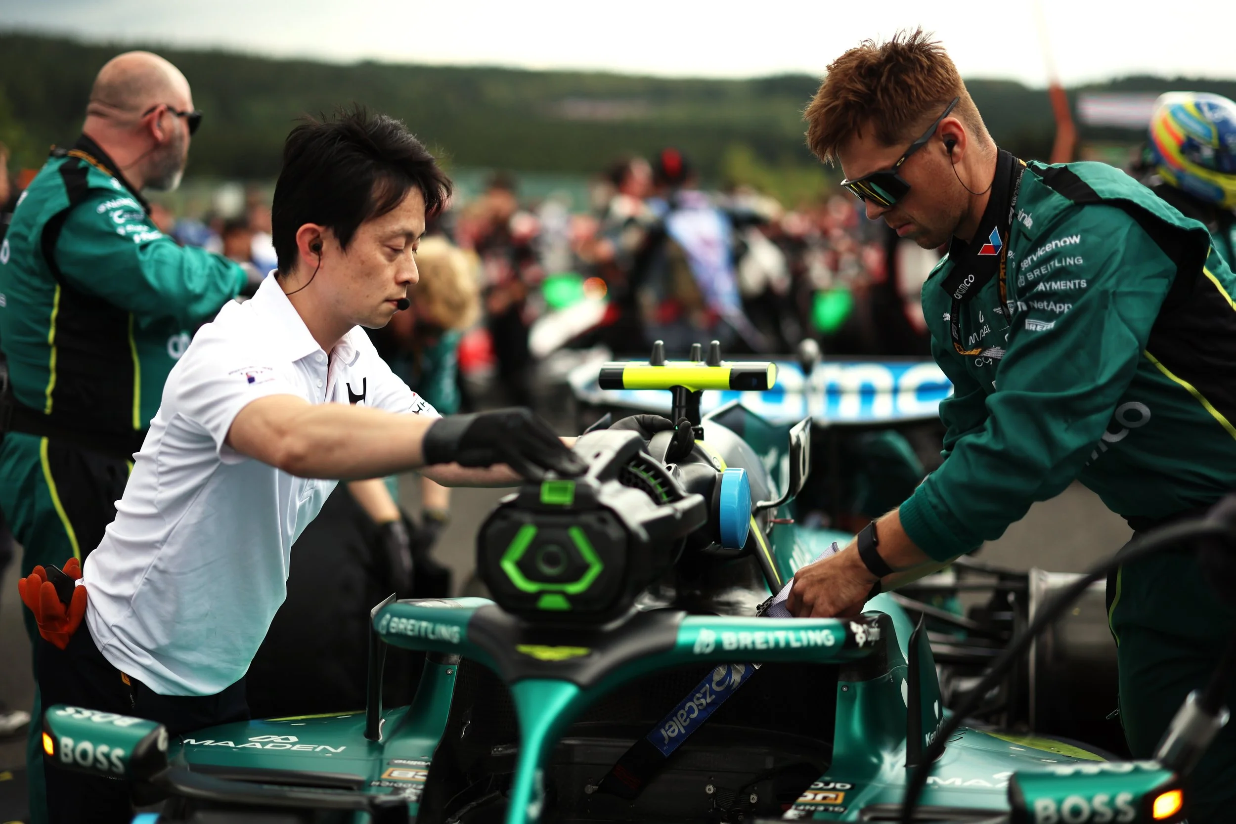 Three people working on a race car in a paddock at a racing event. One man and one woman wearing green team uniforms, and a woman in a white shirt. The background shows a crowd of spectators.