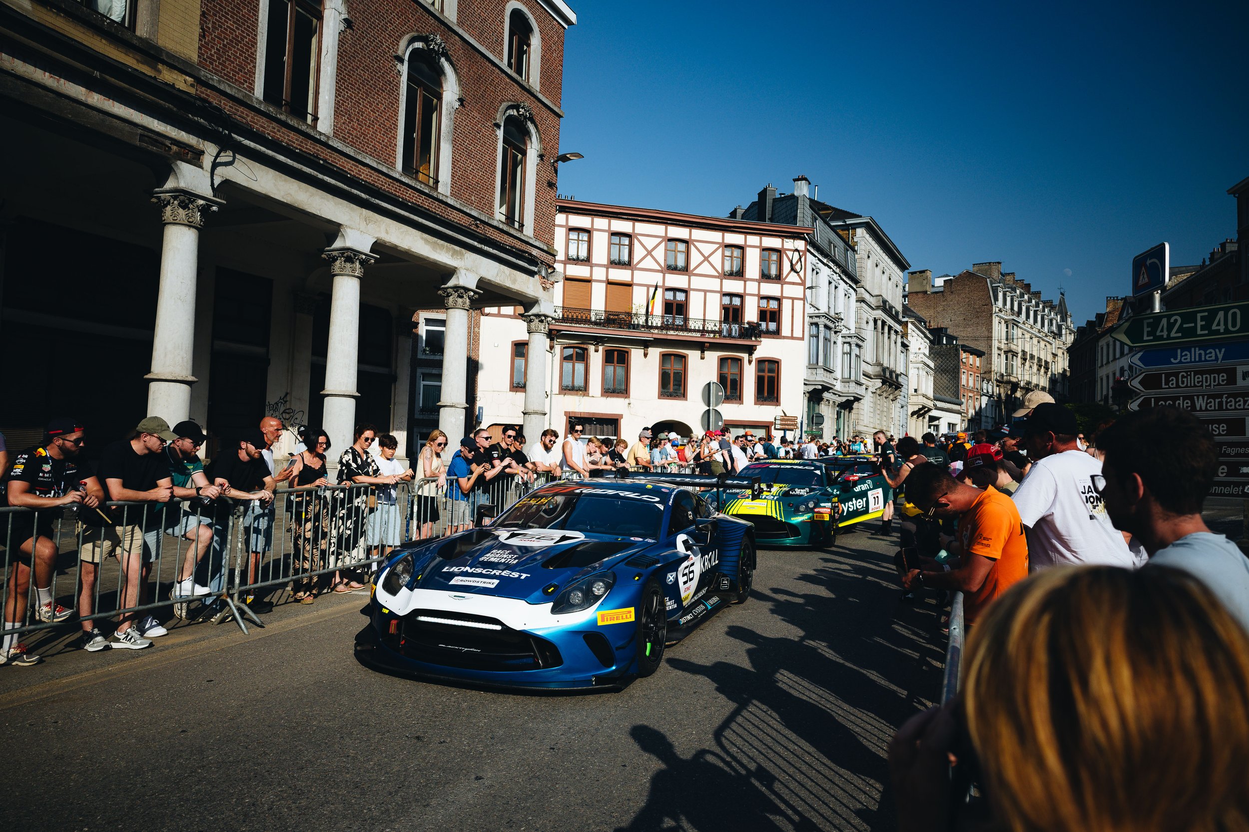 A blue race car with the number 56 on the side, driving down a street lined with spectators behind barriers, with colorful historic buildings in the background under a clear blue sky.