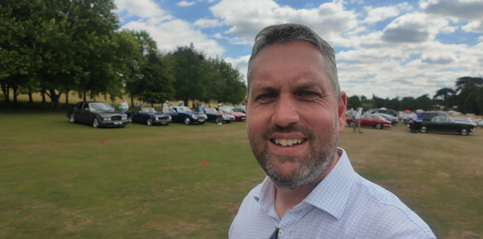 A man taking a selfie outdoors at a classic car show, with vintage cars lined up on a grassy field and trees in the background.