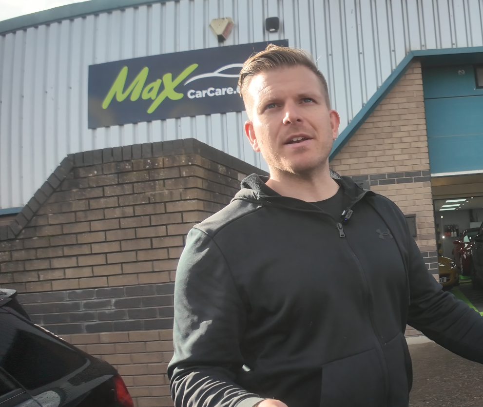 A man with short hair wearing a black jacket standing outside of Max Car Care auto shop.
