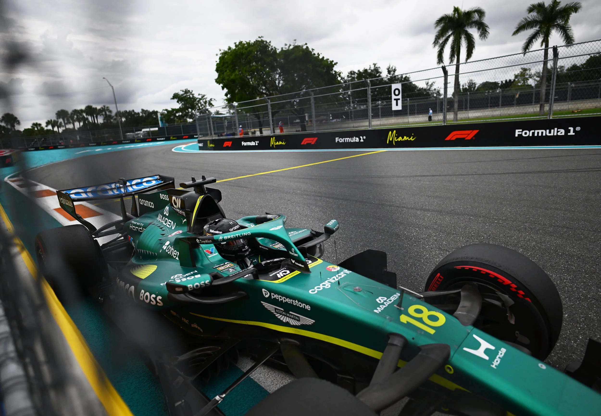Inside view of a Formula 1 race car on a track, showing the steering wheel and cockpit, with a wet racing circuit and trees with palm trees in the background under a cloudy sky.
