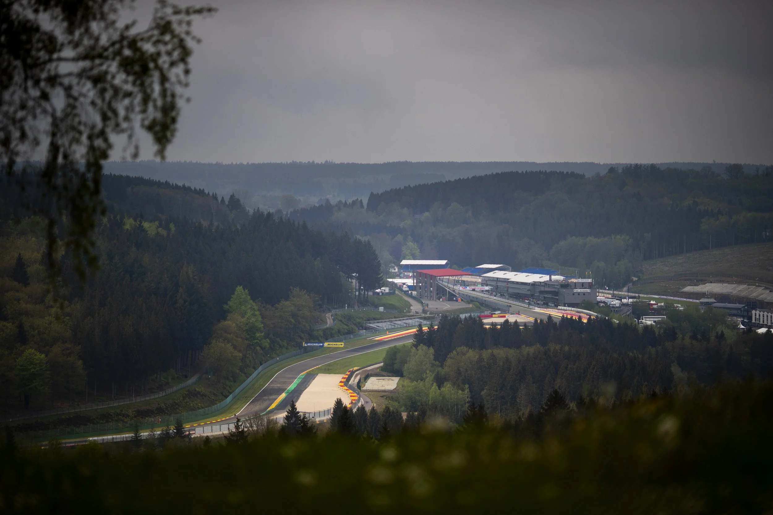 View of Spa race track in a mountainous area with dense trees, overcast sky, and a grandstand with red, white, and blue roofs in the distance.