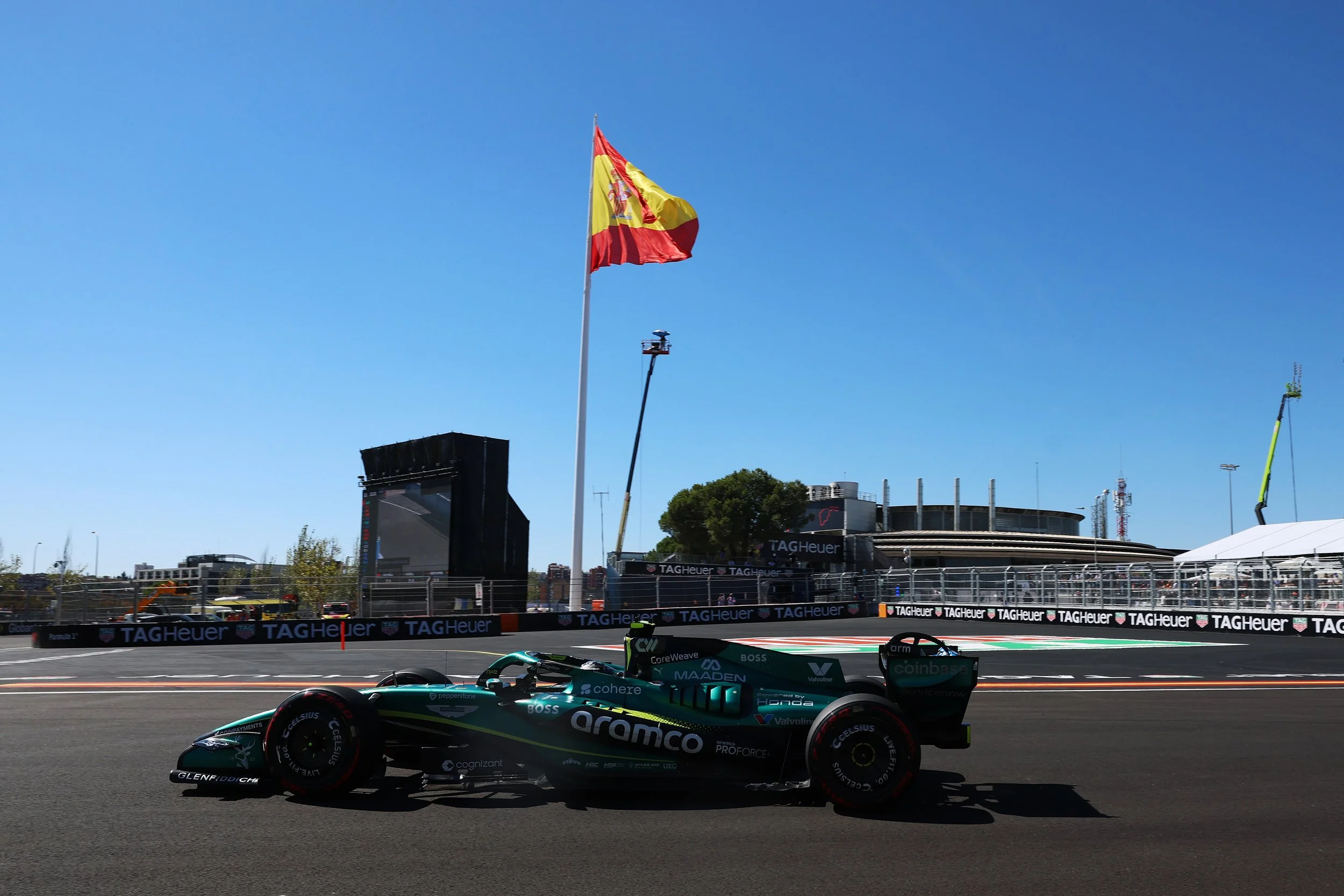 A Formula 1 race car on a race track with a large red and yellow flag flying high in the background against a clear blue sky. The track has sponsor banners, and there are grandstands and buildings in the distance.