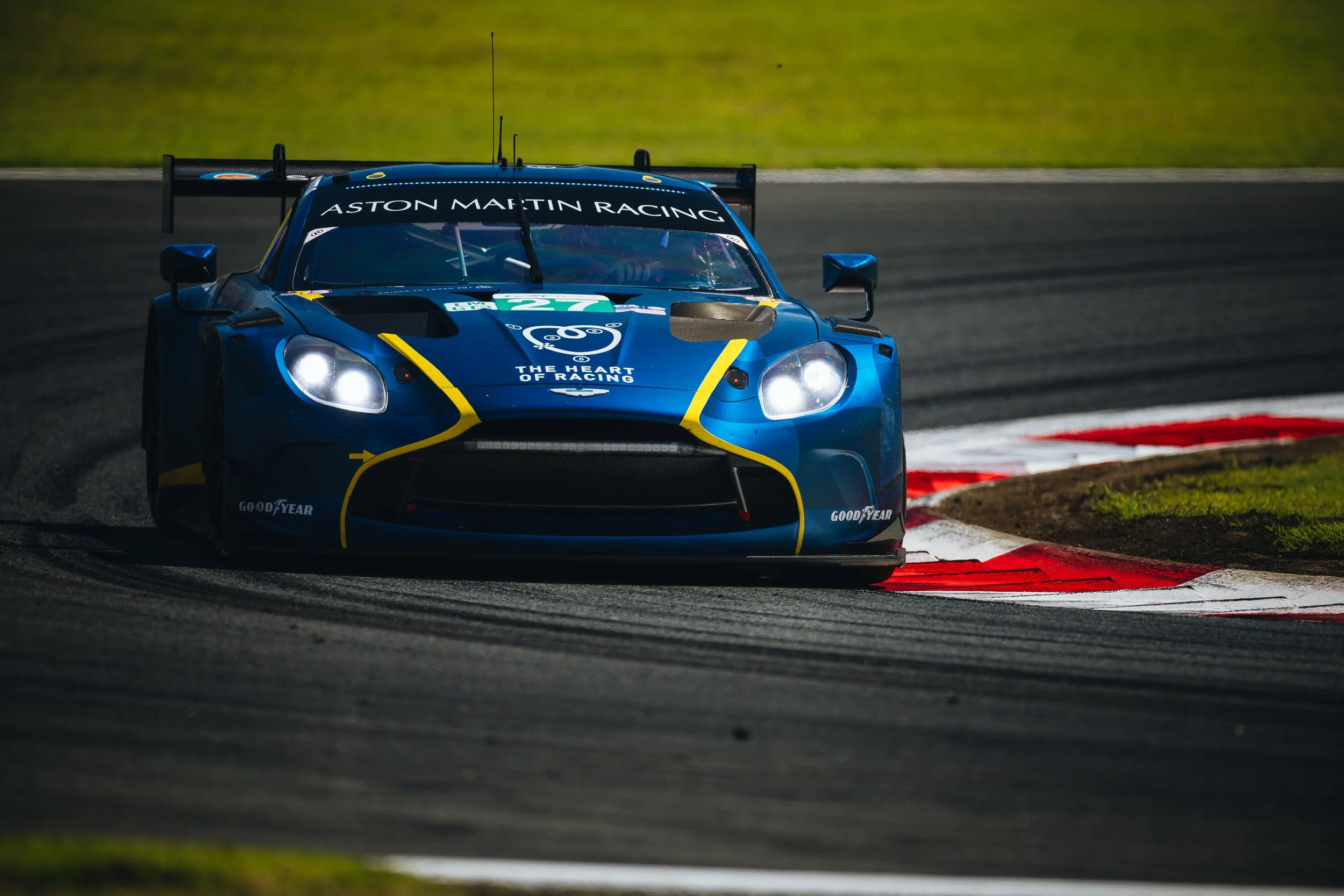 A blue Aston Martin racing car with the logo 'The Heart of Racing' and number 27, on a race track with red and white curbs, taking a turn during a race.