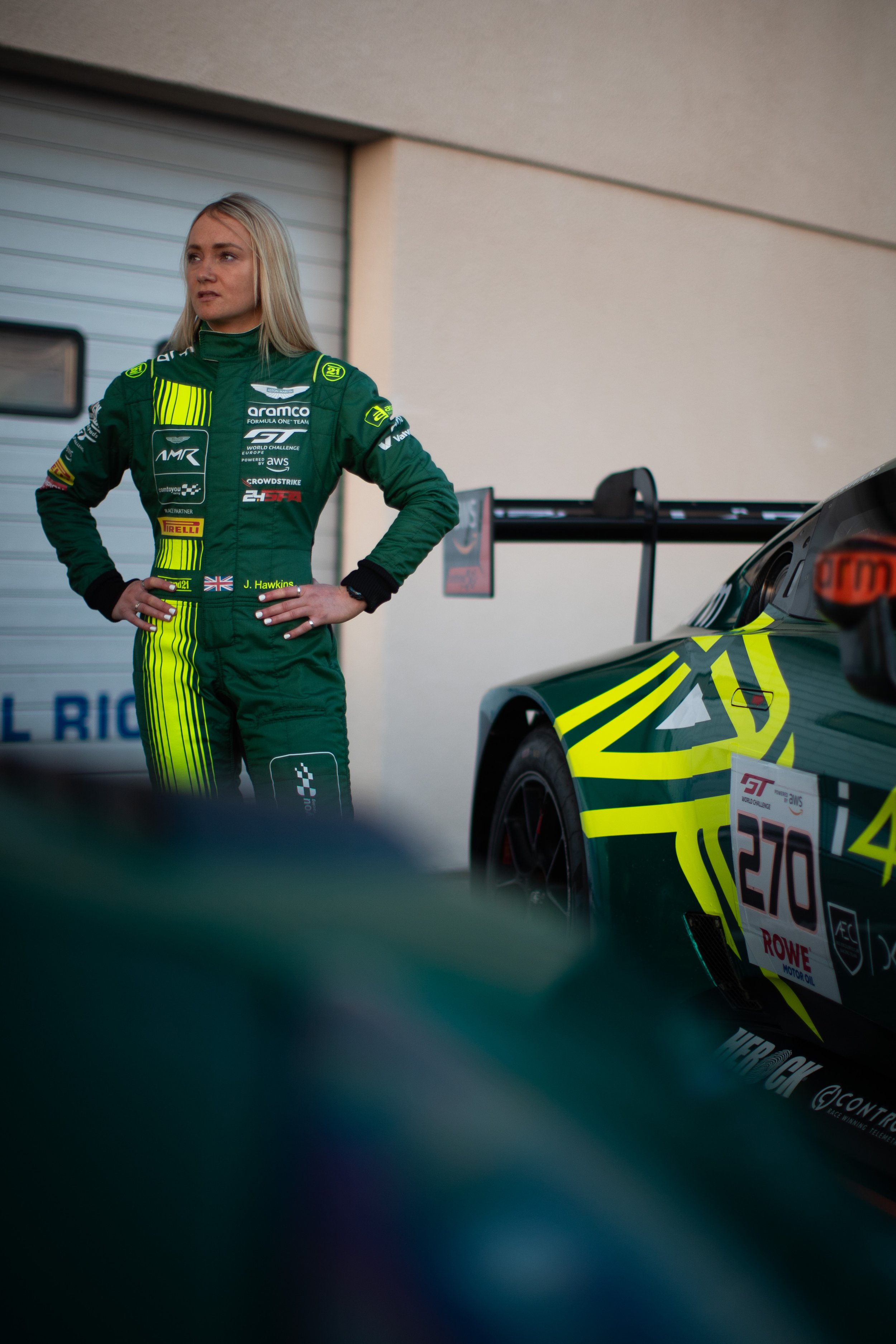 A woman in a green racing suit with various sponsor logos stands with her hands on her hips, next to a race car with a colorful design, in a garage or paddock area.