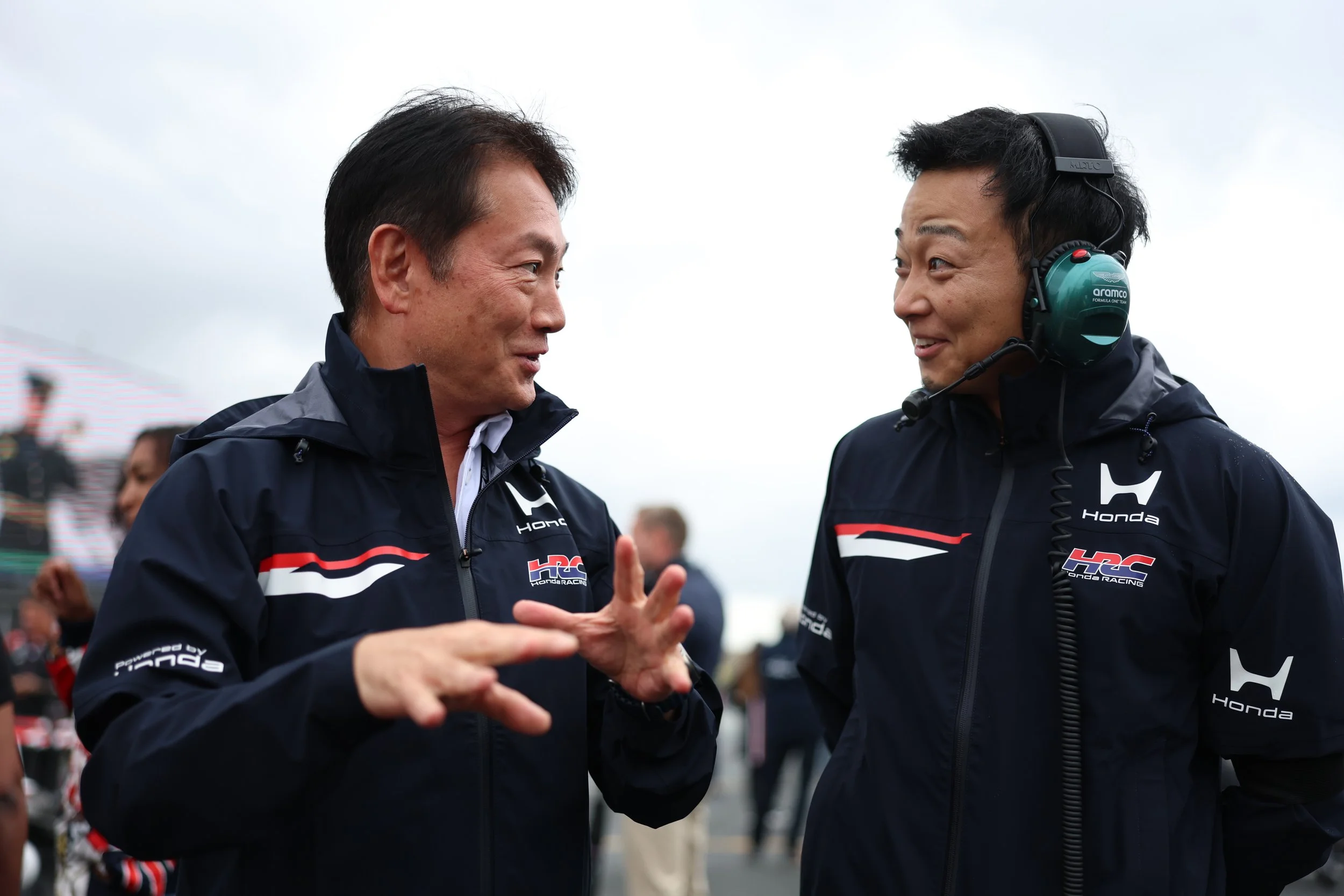 Two men in Honda Racing jackets engaging in conversation at an outdoor motorsport event, with a crowd and racing crew in the background under cloudy skies.