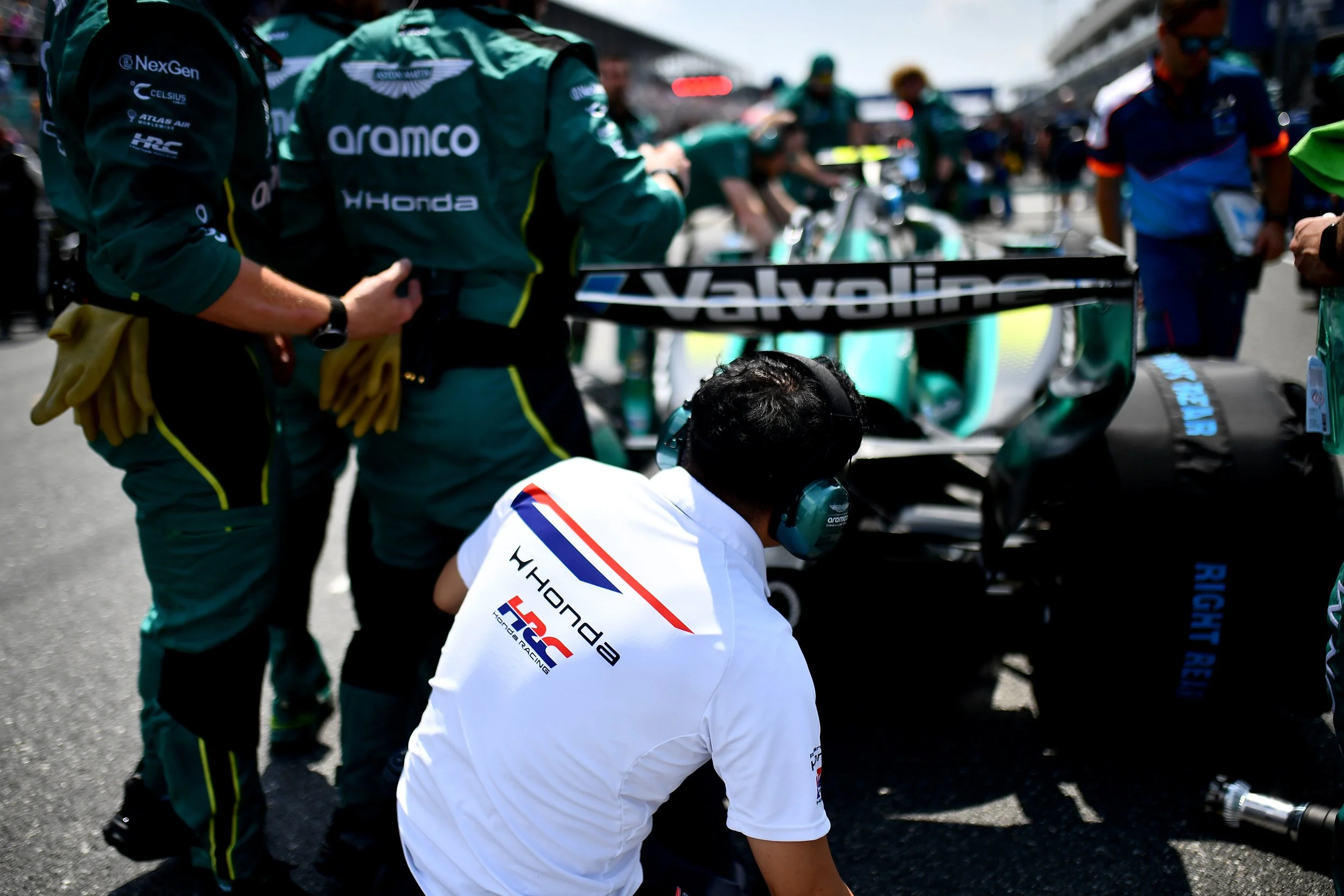 Race car team members in green and blue uniforms preparing at a racing event, with racing cars in the background.