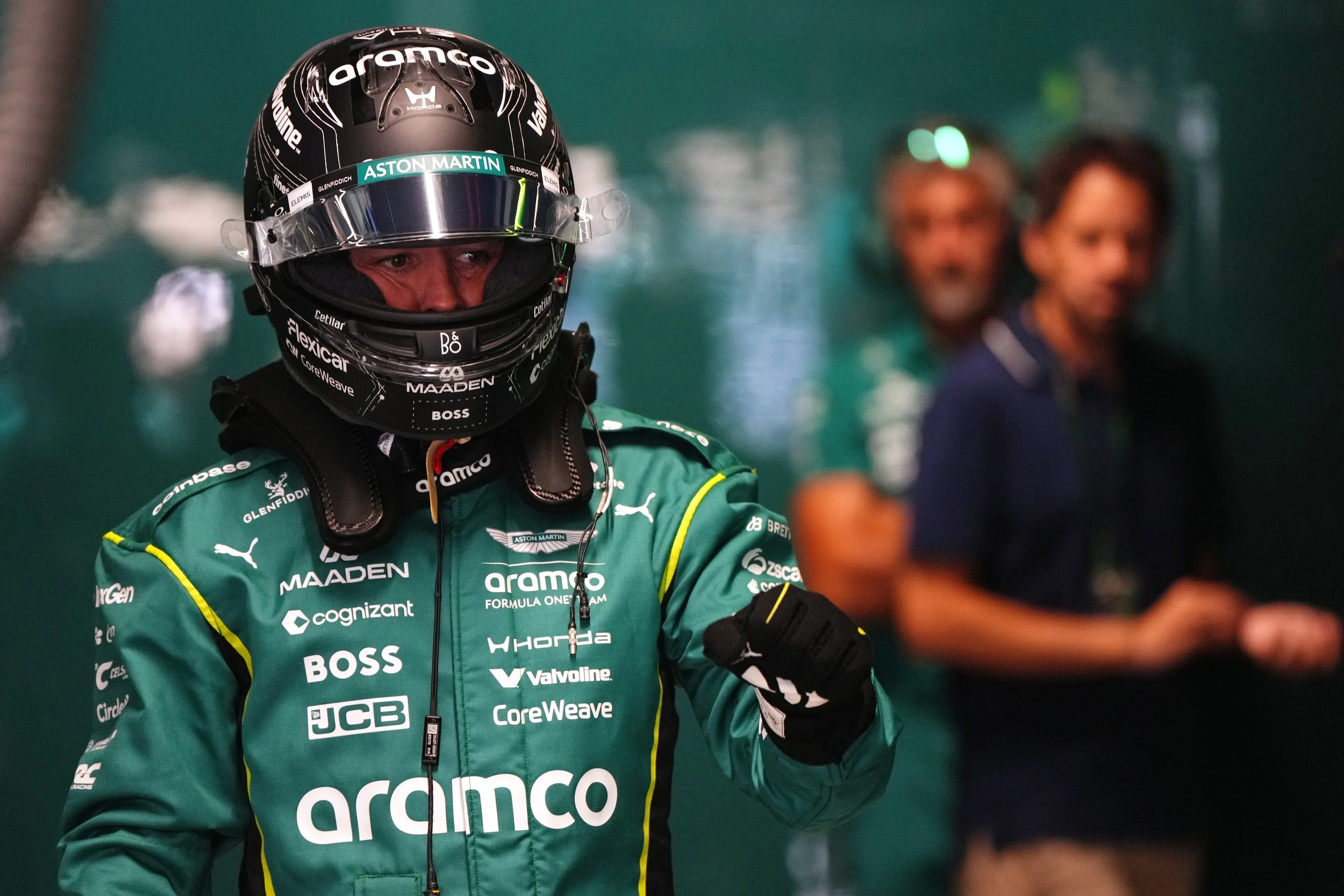 A race car driver in a green racing suit and black helmet with a reflective visor, featuring sponsorship logos. Two out-of-focus individuals are in the background.