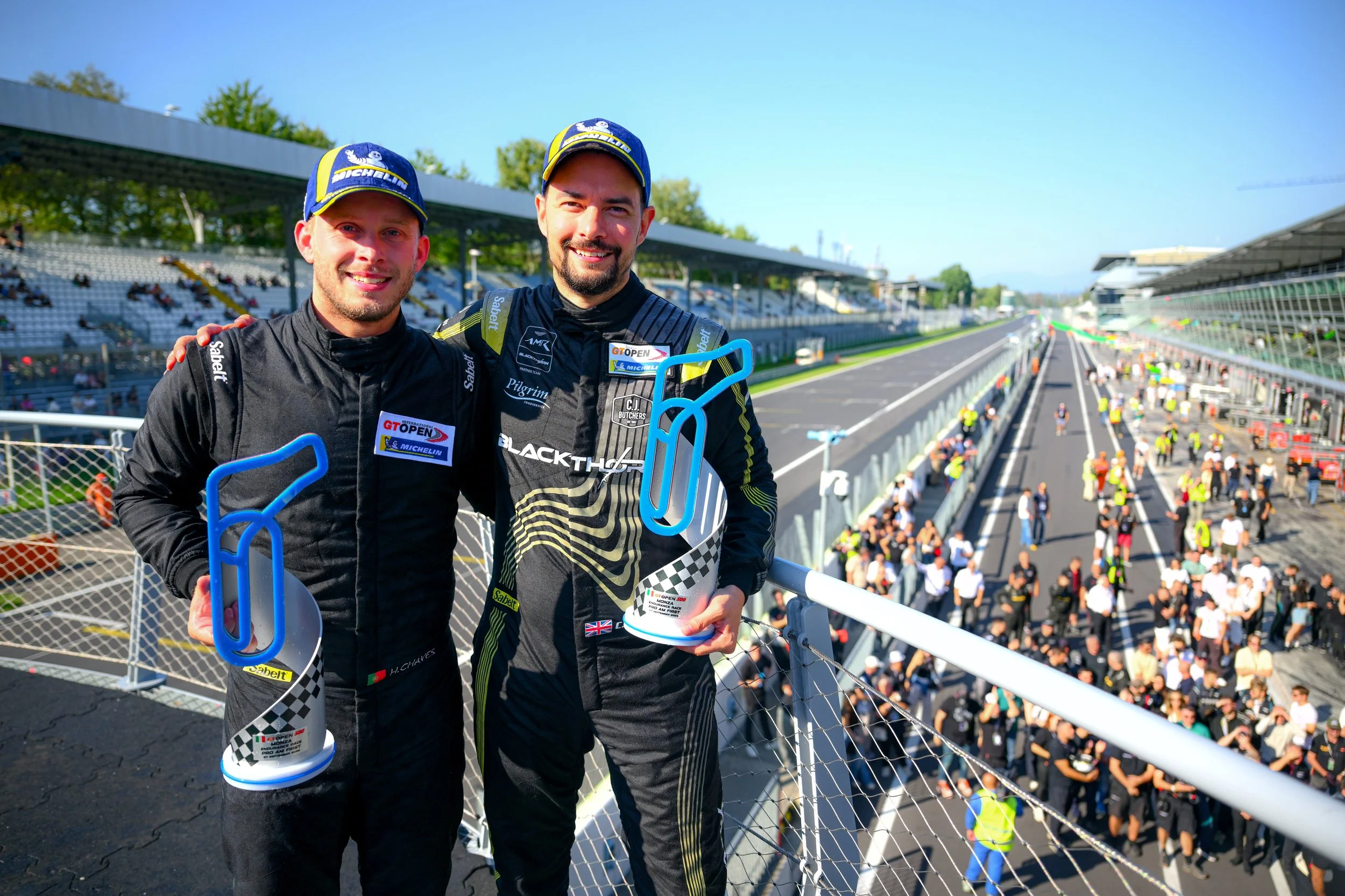 Two race car drivers in black racing suits and blue caps smiling while holding awards on a race track grandstand.