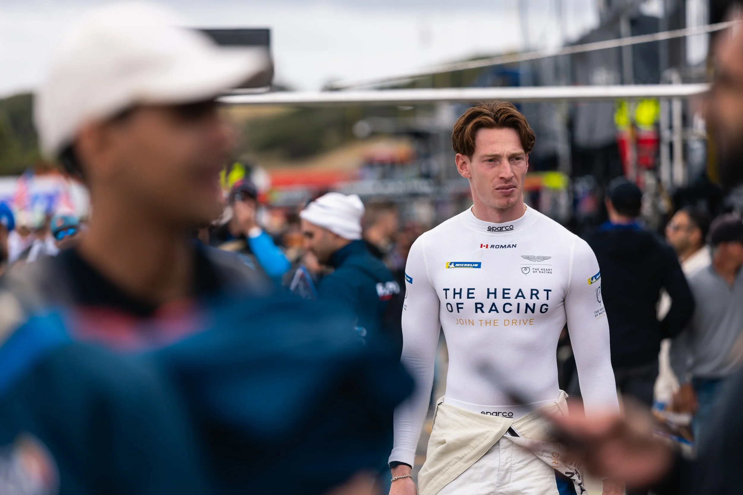 A young man in a white racing suit with sponsorship logos, including 'The Heart of Racing,' stands in a racing paddock area surrounded by people. He appears focused, with reddish-brown hair, and is looking slightly to the right. The background is slightly blurred, with various individuals and equipment, indicating a motorsport event.