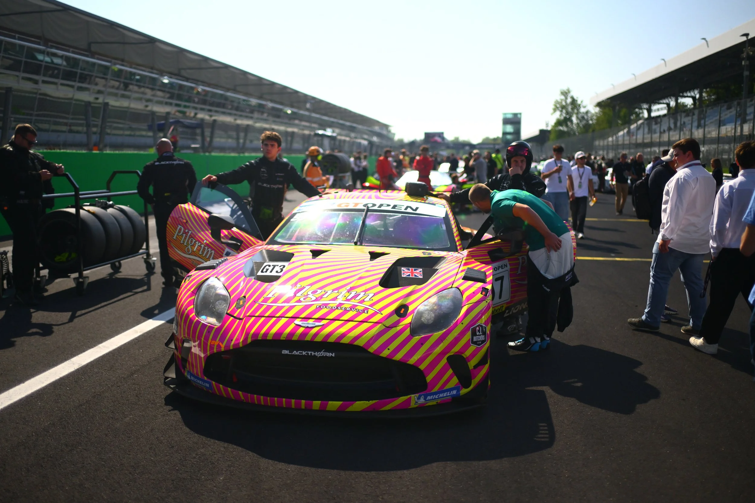 A pink and yellow race car with various sponsorship stickers and the number 7. People, including team members and officials, surround the car in a pit lane at a racing event.