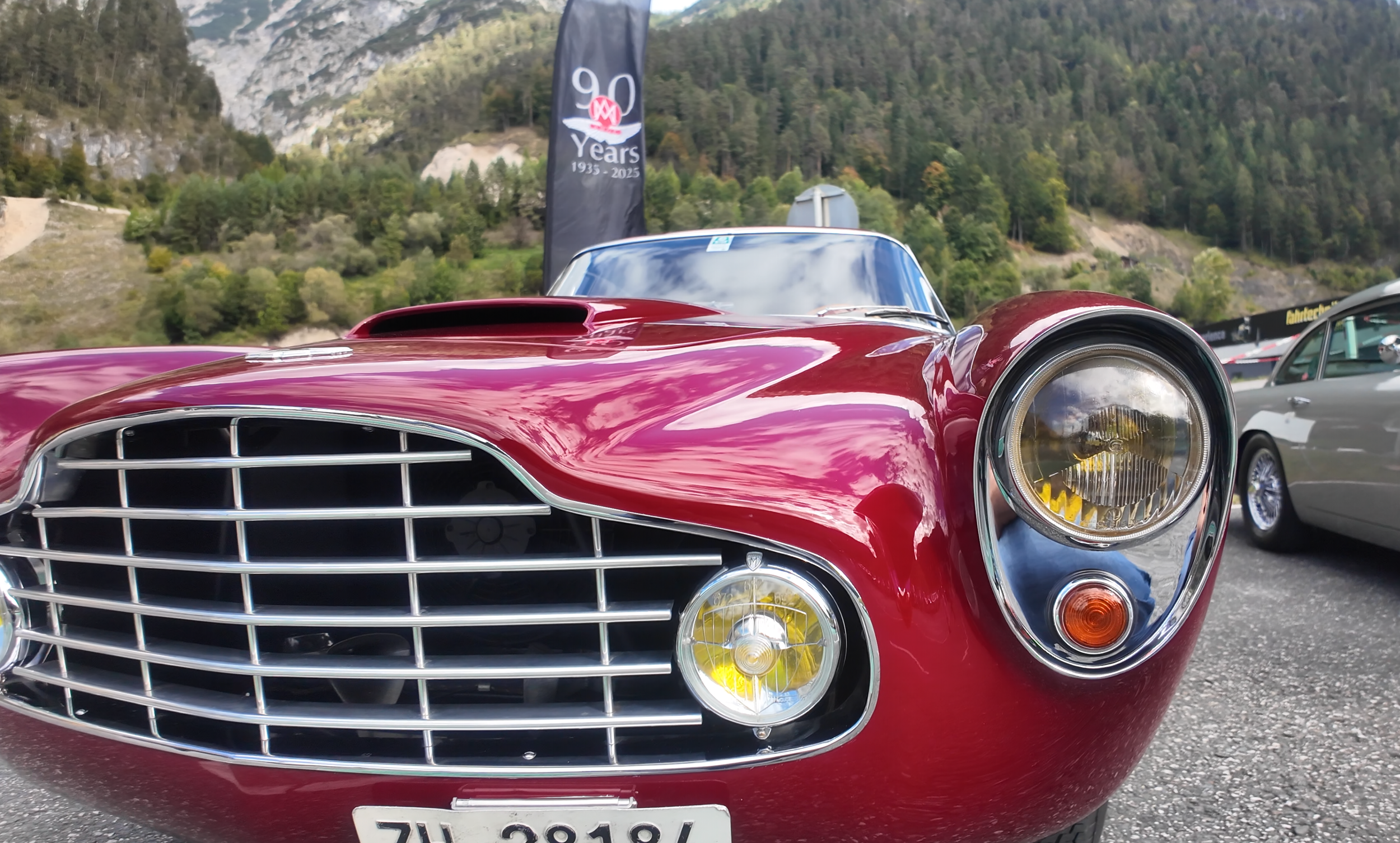 Close-up of a vintage red Aston Martin sports car at a car show, with a prominent front grille, headlights, and a mountain landscape in the background. A banner celebrating 90 years is visible.