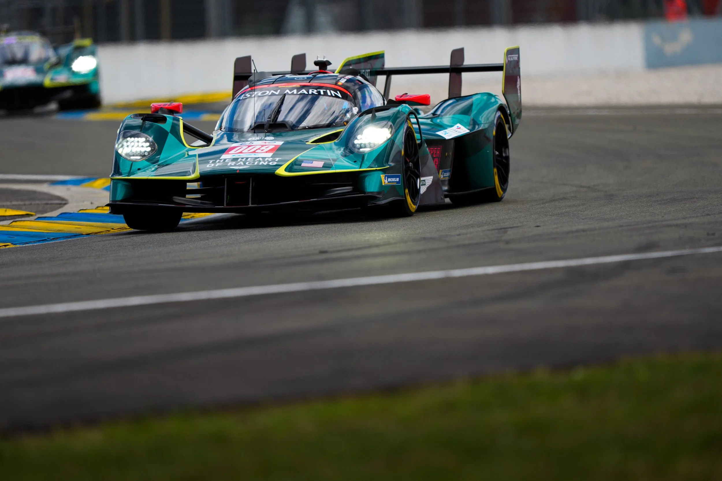 A race car on a race track with another car blurred in the background, featuring sponsor logos and a sleek aerodynamic design.
