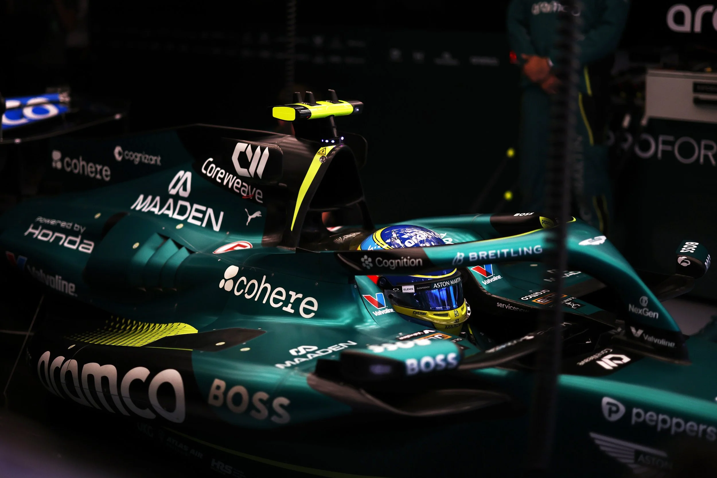 Close-up of a Formula 1 race car with sponsorship logos, including 'cohere,' 'MADDEN,' 'BOSS,' and 'peppers.' The driver in the cockpit is wearing a helmet with blue and yellow design. The night setting highlights the car's sleek design and branding.