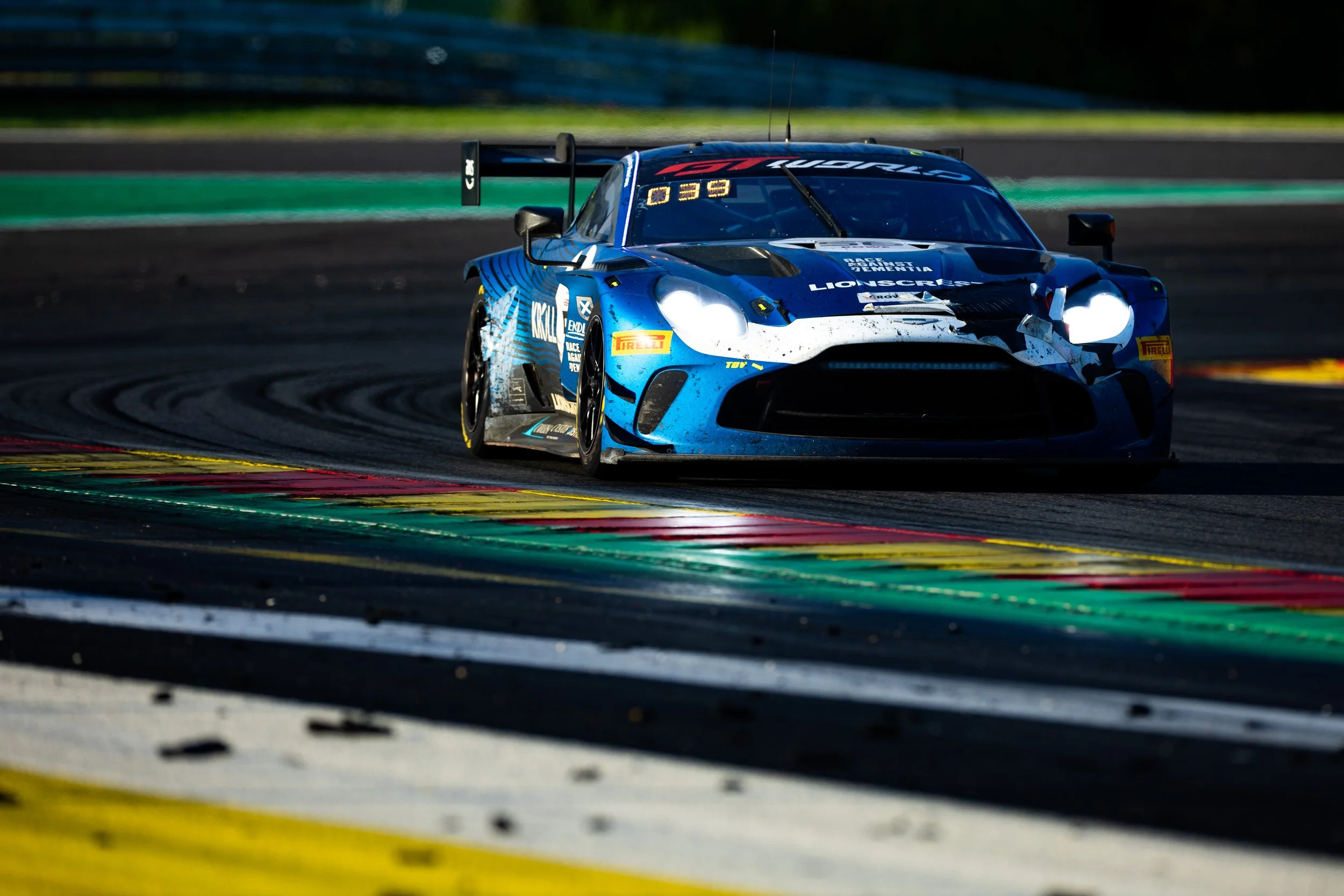 A blue race car speeding on a race track, kicking up dirt, with a large rear wing and various sponsor decals, during a race.