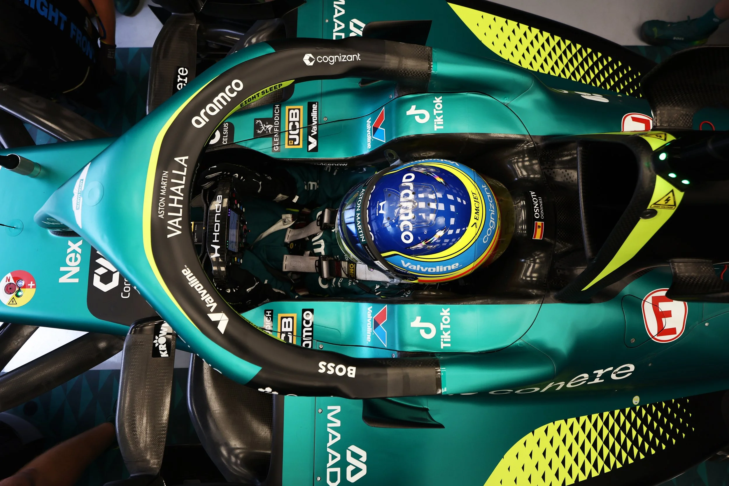 Fernando Alonso, F1 Driver for Aston Martin in his race car seated in a teal and black racing car in the garage, wearing a blue helmet with yellow and green accents, and a black racing suit with sponsors' logos.