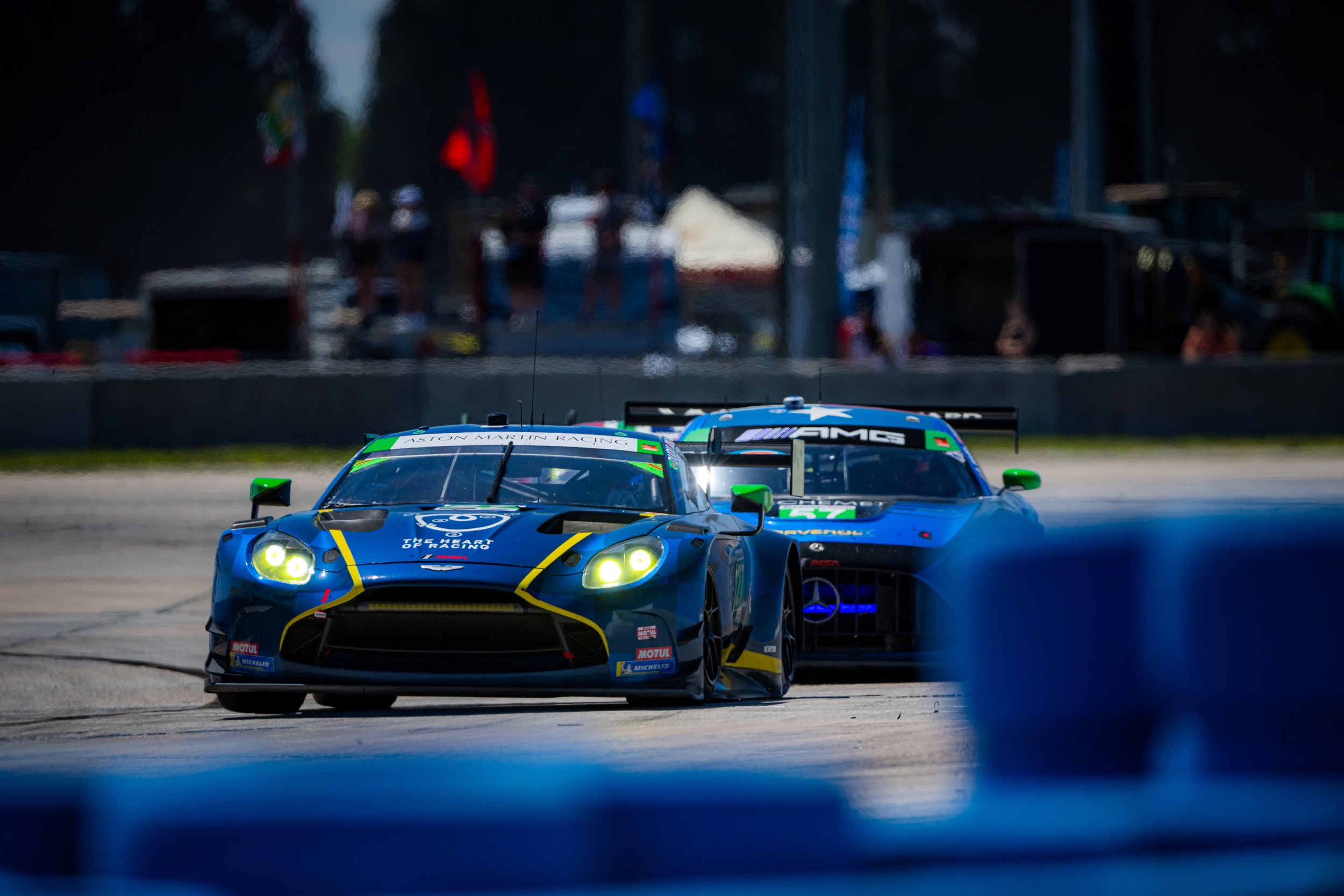 Two blue race cars with yellow accents driving on a race track, with pit area and people in the background.