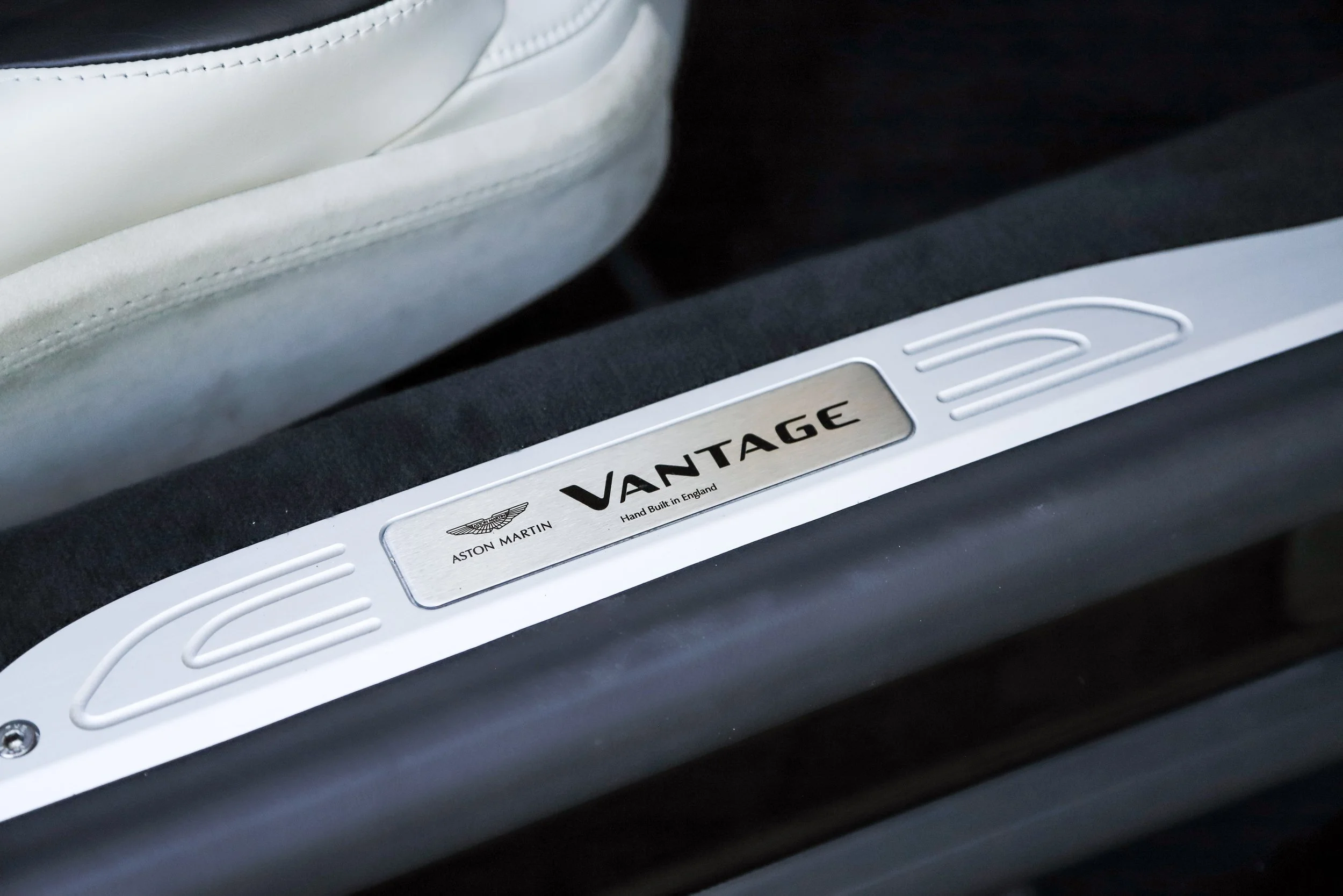 Close-up of a metal door sill plate inside a car with 'VANTAGE' written on it, along with the Aston Martin logo and text indicating it is hand-built in England, next to a beige car seat and black floor mat.