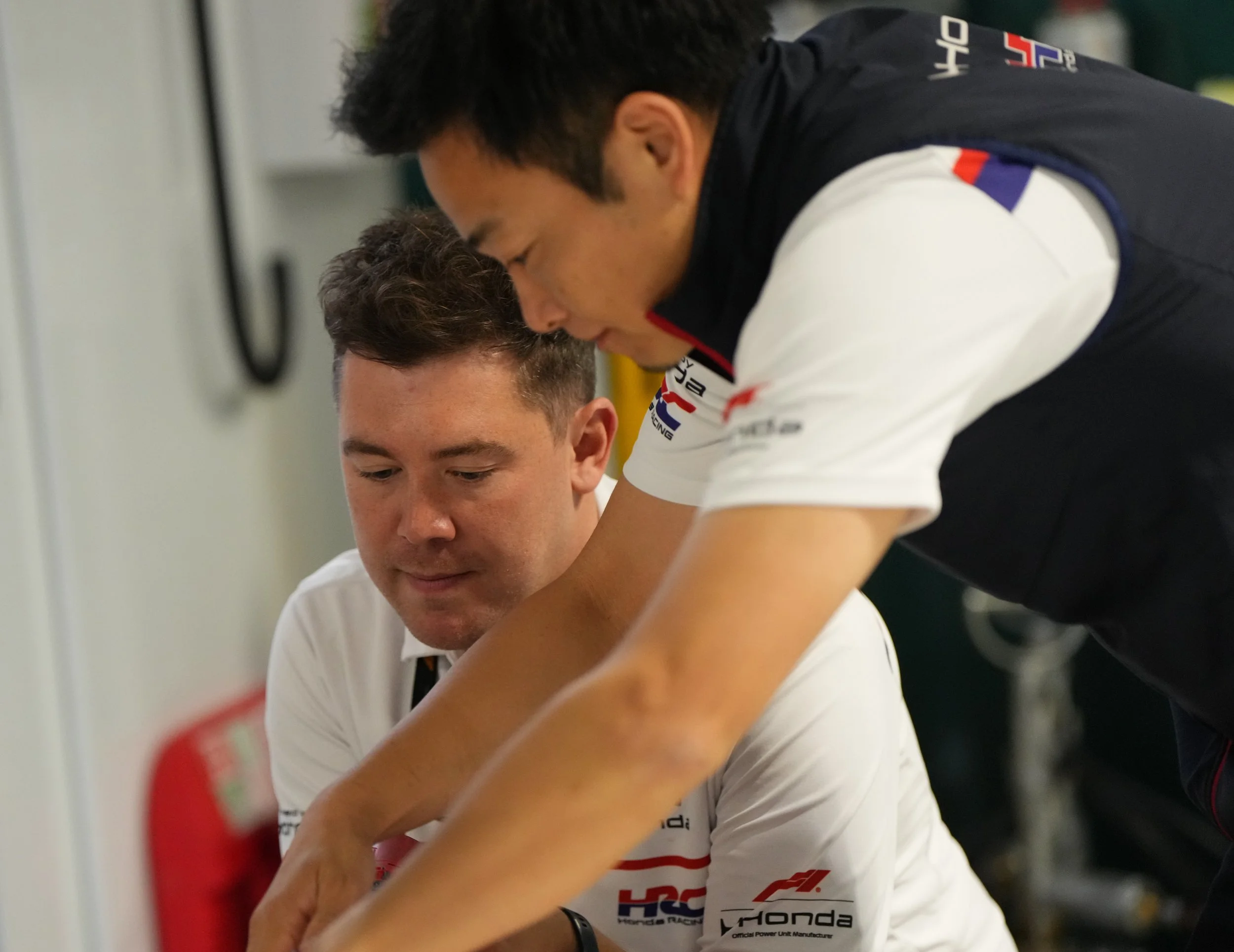 Two men, one in a black vest and another in a white shirt, are leaning close together, engaging in a focused conversation or activity, in a room with a white wall and a blurred red object in the background.