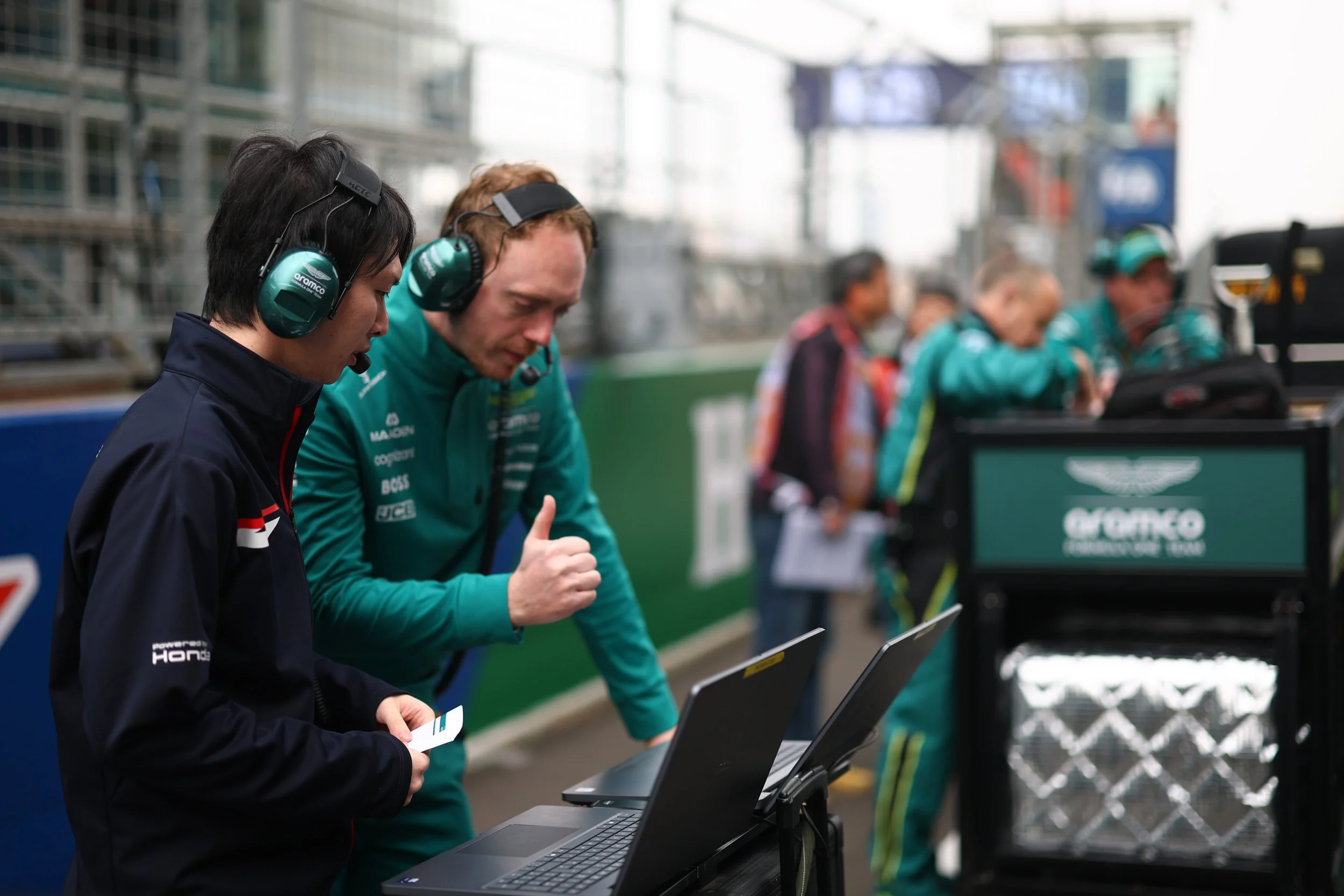 Formula 1 engineers working at a race track, wearing headsets and looking at laptops.