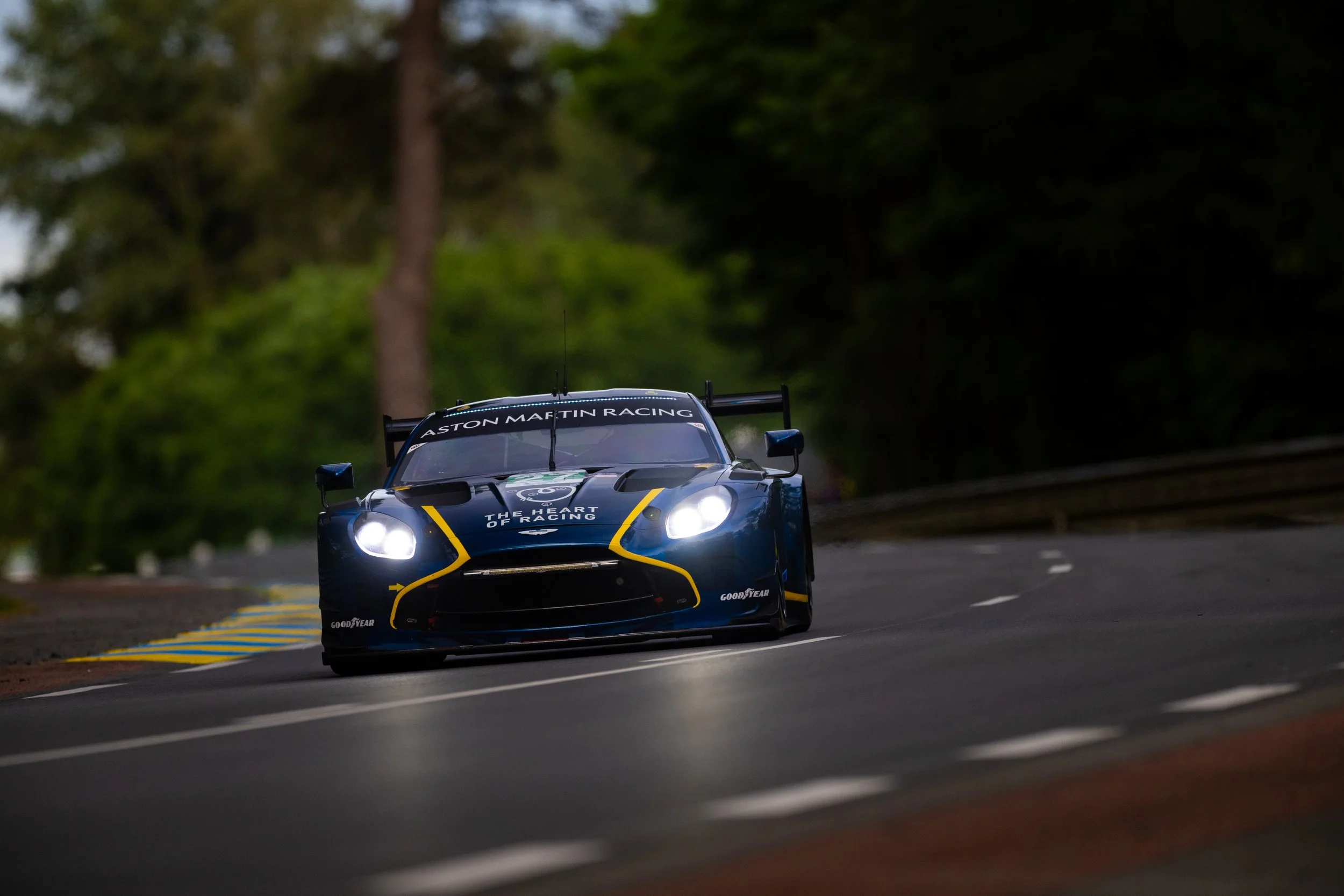 A blue Aston Martin race car with yellow accents driving on a curved road surrounded by green trees during the daytime.