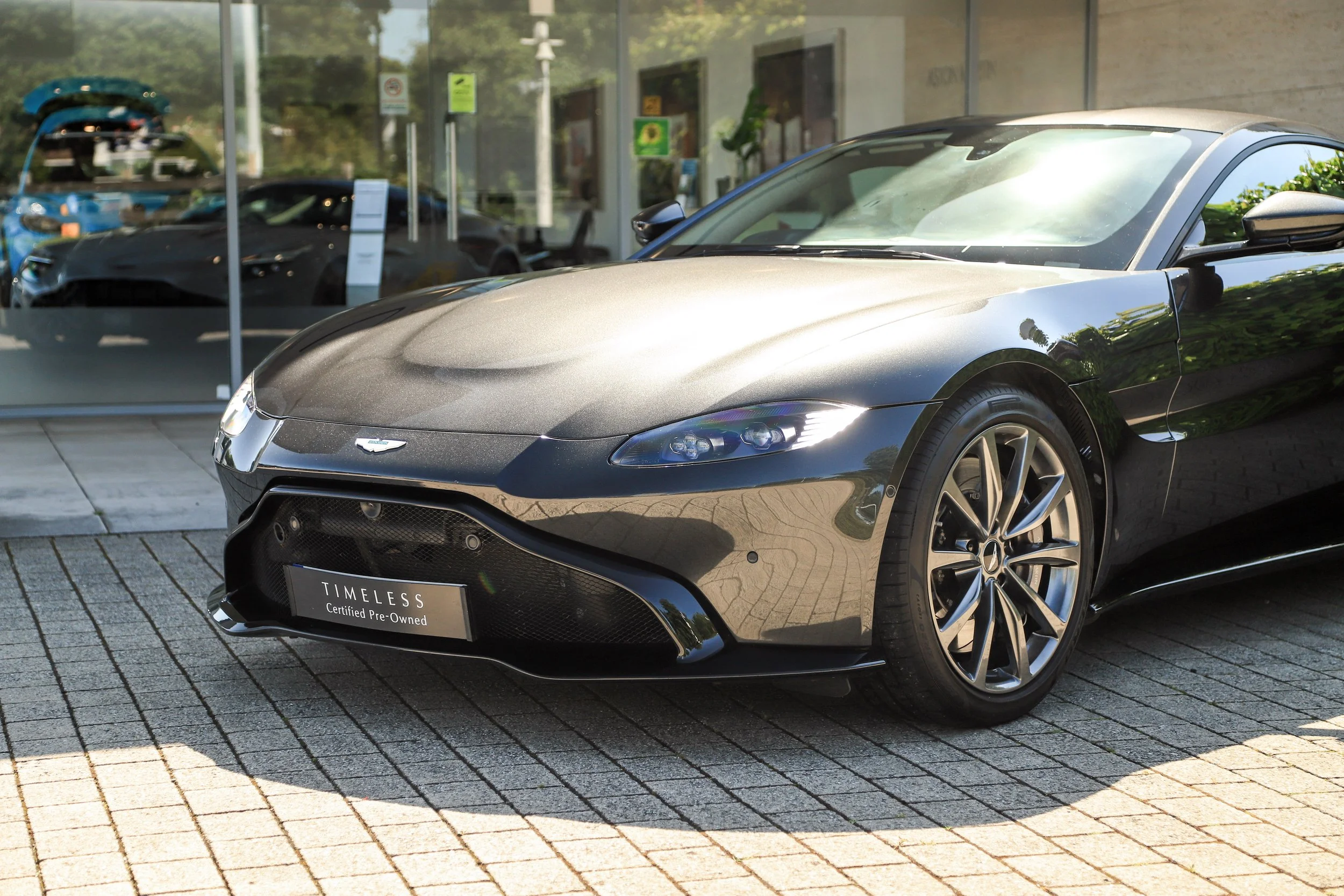 A black Aston Martin sports car parked outside a dealership with a glass storefront, cars visible inside, and a sign that reads "TIMELESS Certified Pre-Owned."