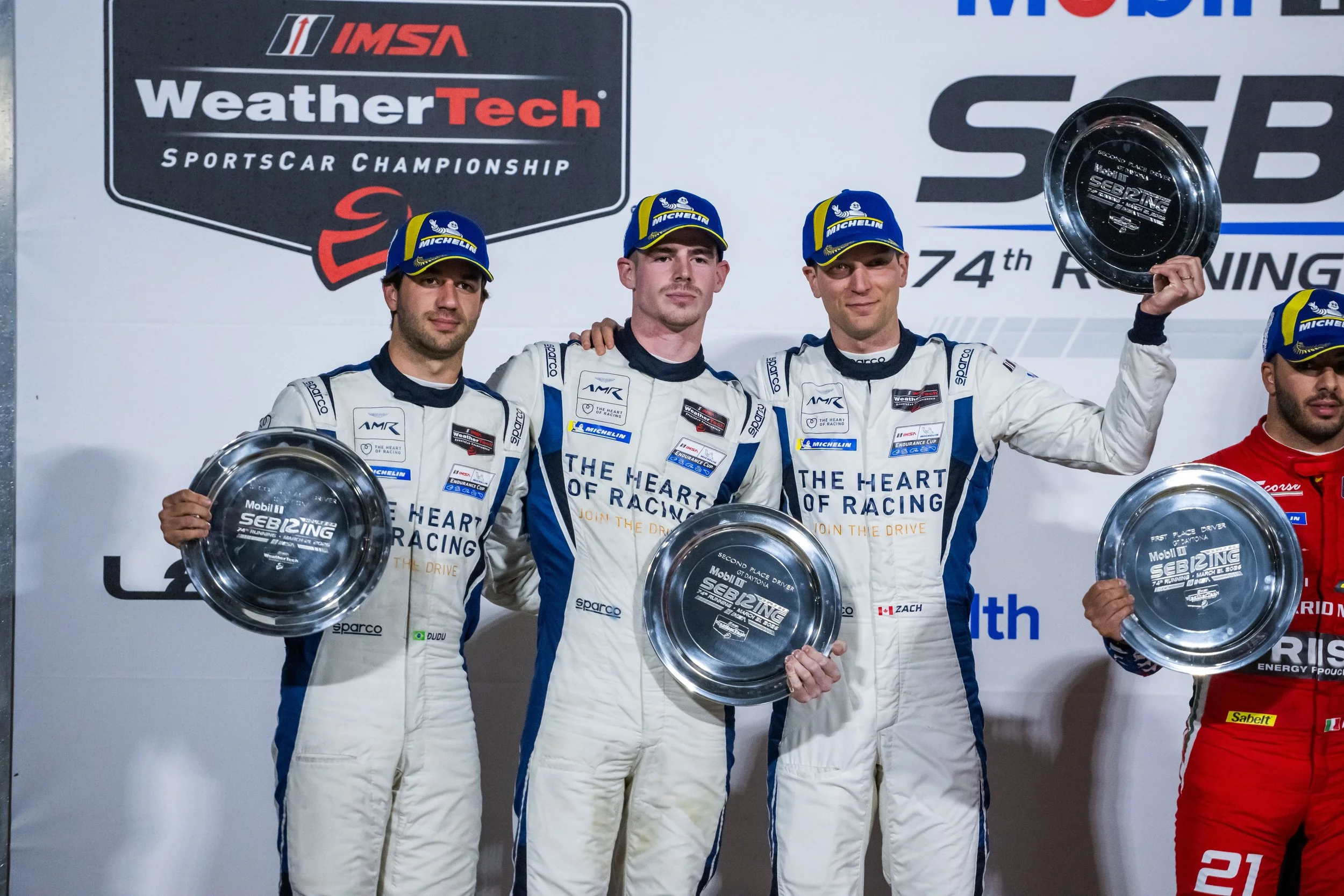 Three race car drivers in white racing suits and blue caps with Michelin logos holding silver plates on the podium. They are standing in front of a large racing event banner that reads WeatherTech Sportscar Championship. The middle driver is in first place, and the driver on the left is second. The driver on the right is holding the third-place plate.