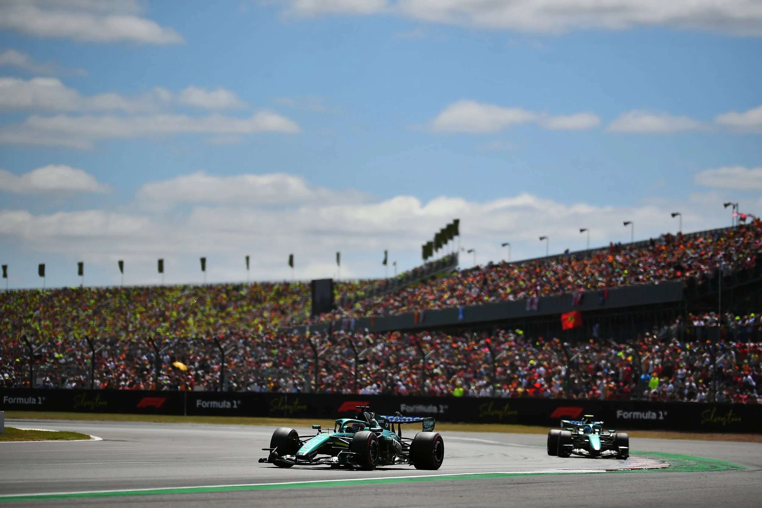 Two Formula 1 race cars on a racetrack with a large crowd in the grandstands and blue sky with clouds overhead.