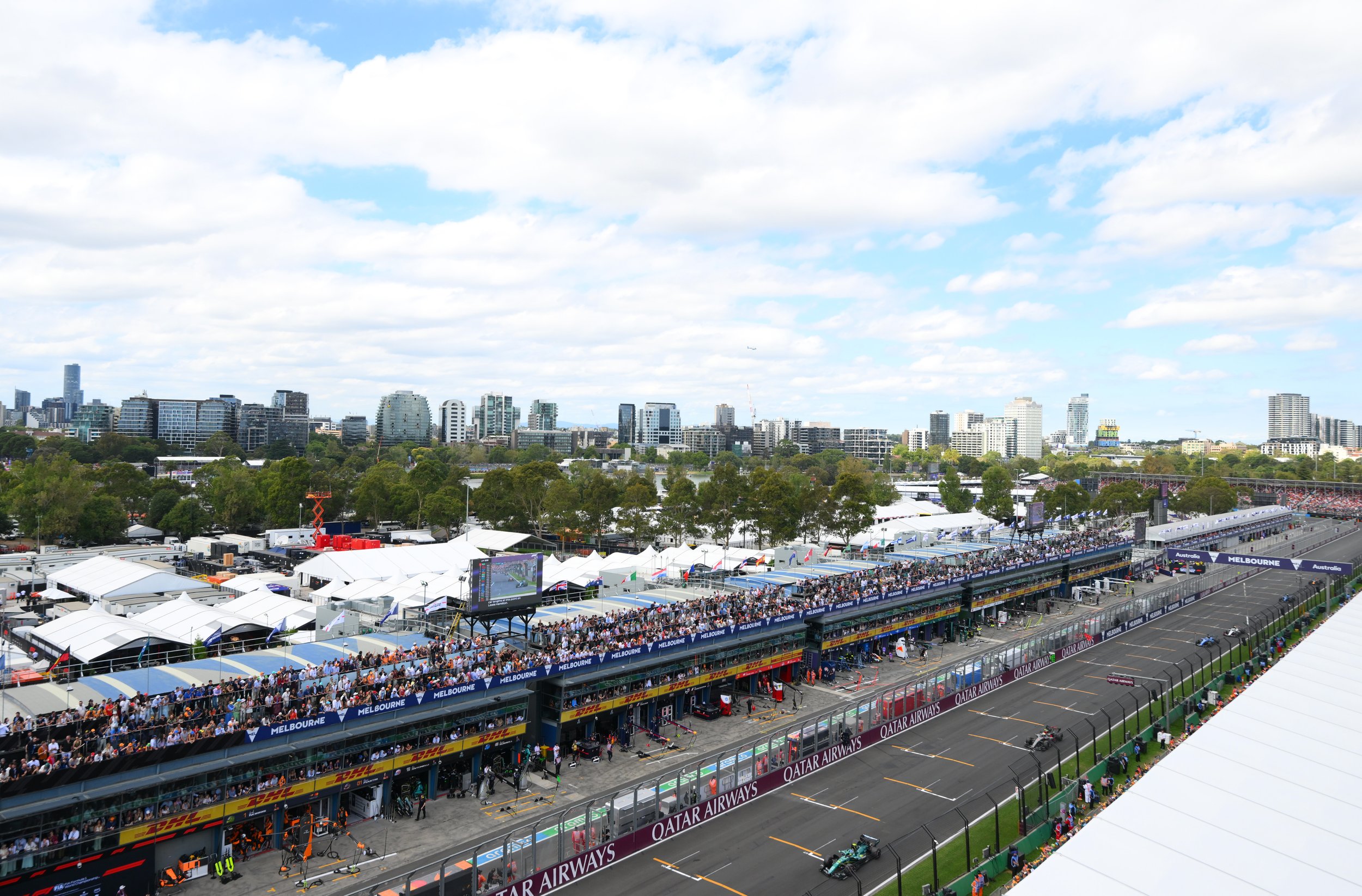 View of a race track with Formula 1 cars on it, a partly cloudy sky, and a busy racing event at the Melbourne Grand Prix circuit with spectators in the grandstands.