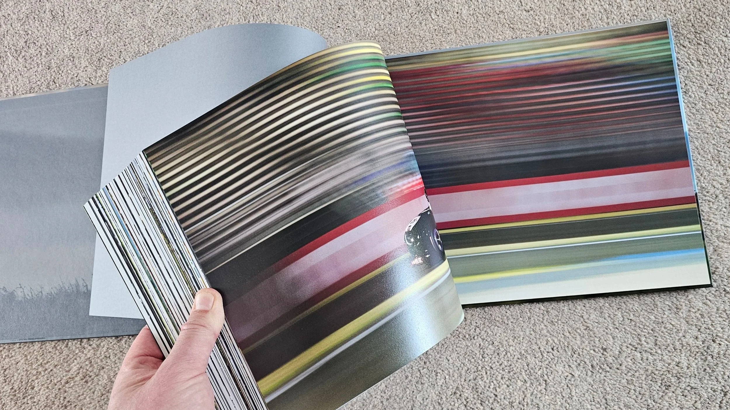 Open book called LM4 'The life story of an Aston Martin' displaying a blurred digital photo of a moving car on a racetrack, with a person's hand holding the page, on a beige carpeted floor.