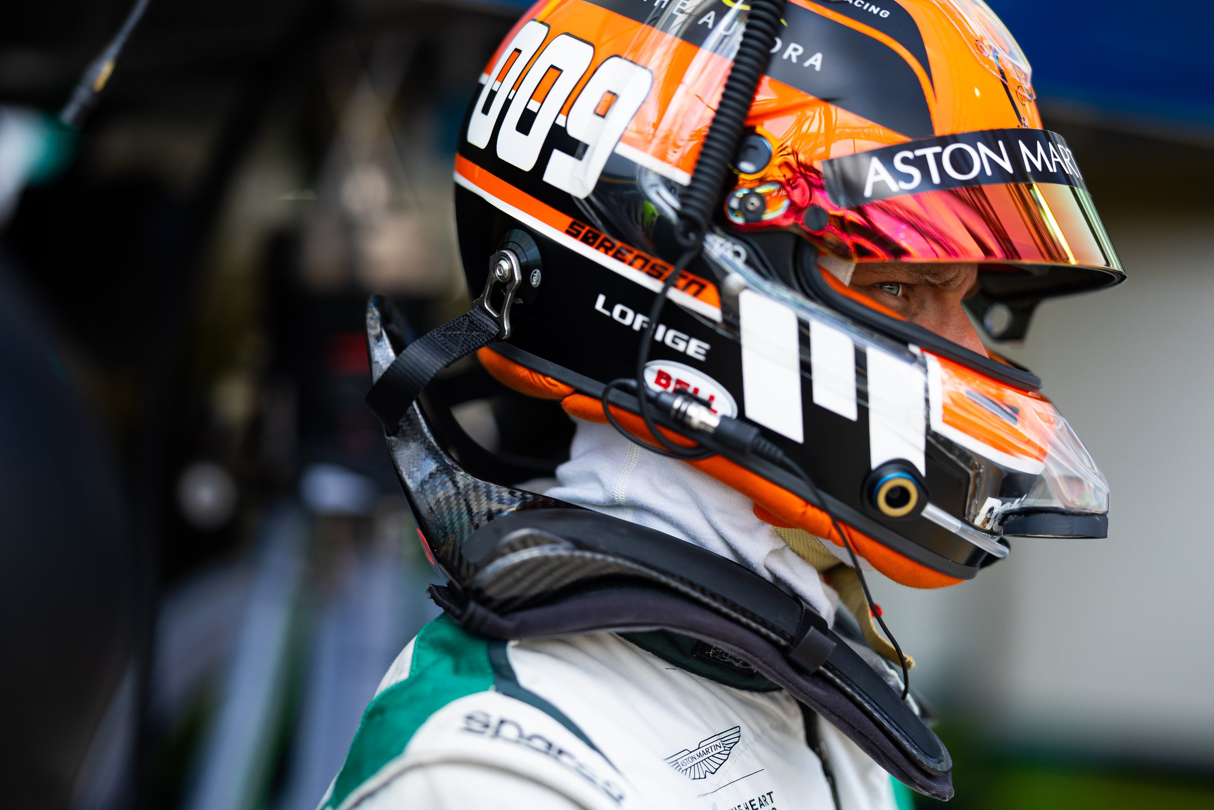 Close-up of a race car driver wearing a white racing suit and an orange helmet with a reflective visor, sitting in the cockpit.