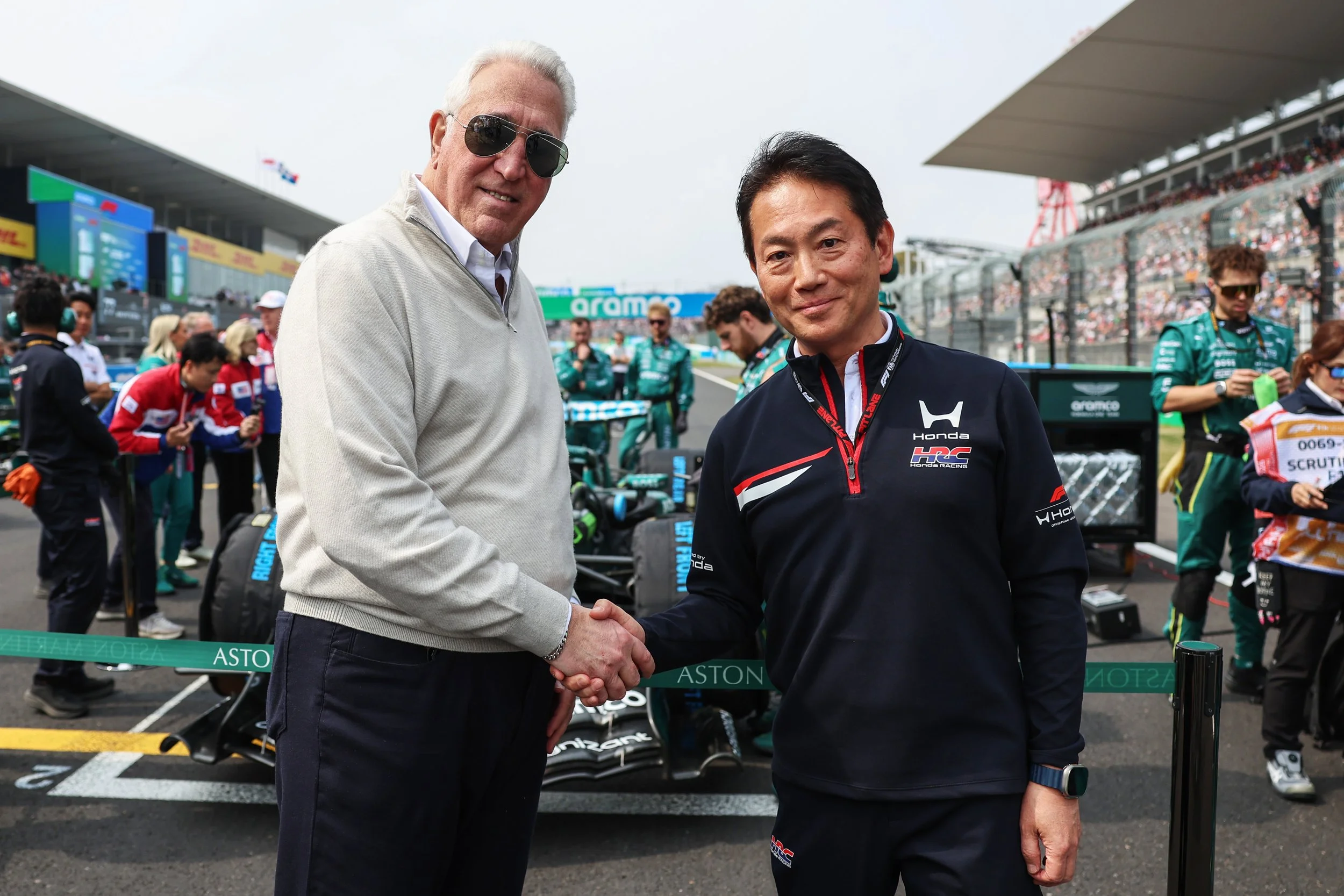 Two men shaking hands at a race track, with racing teams and cars in the background. One man is wearing sunglasses and a light-colored sweater, and the other is in a black Honda racing team jacket.
