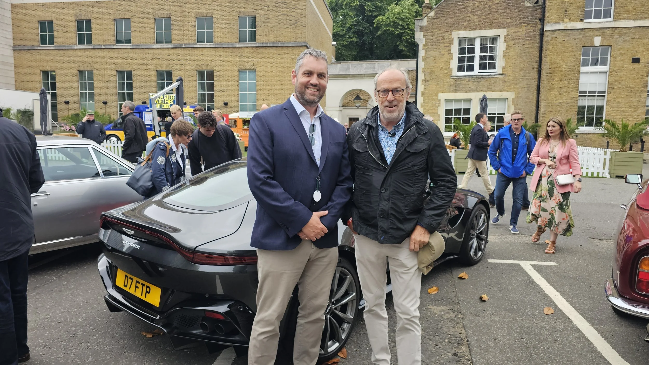 Two men standing in front of a black Aston Martin car at an outdoor car event. The man on the left has gray hair, a beard, and is wearing a blue blazer and khaki pants. The man on the right has glasses, gray hair, and is wearing a black jacket and beige pants. Other people and vintage cars are visible in the background.