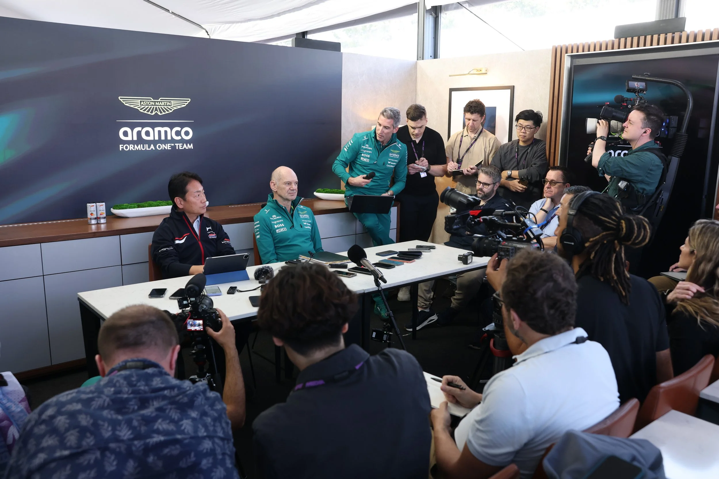 Members of the Aston Martin Formula One team are at a press conference or briefing, with several journalists and media personnel capturing the event on cameras. The backdrop displays the Aston Martin logo and the words "Aramco Formula One Team." The room is bright with natural light coming through large windows.