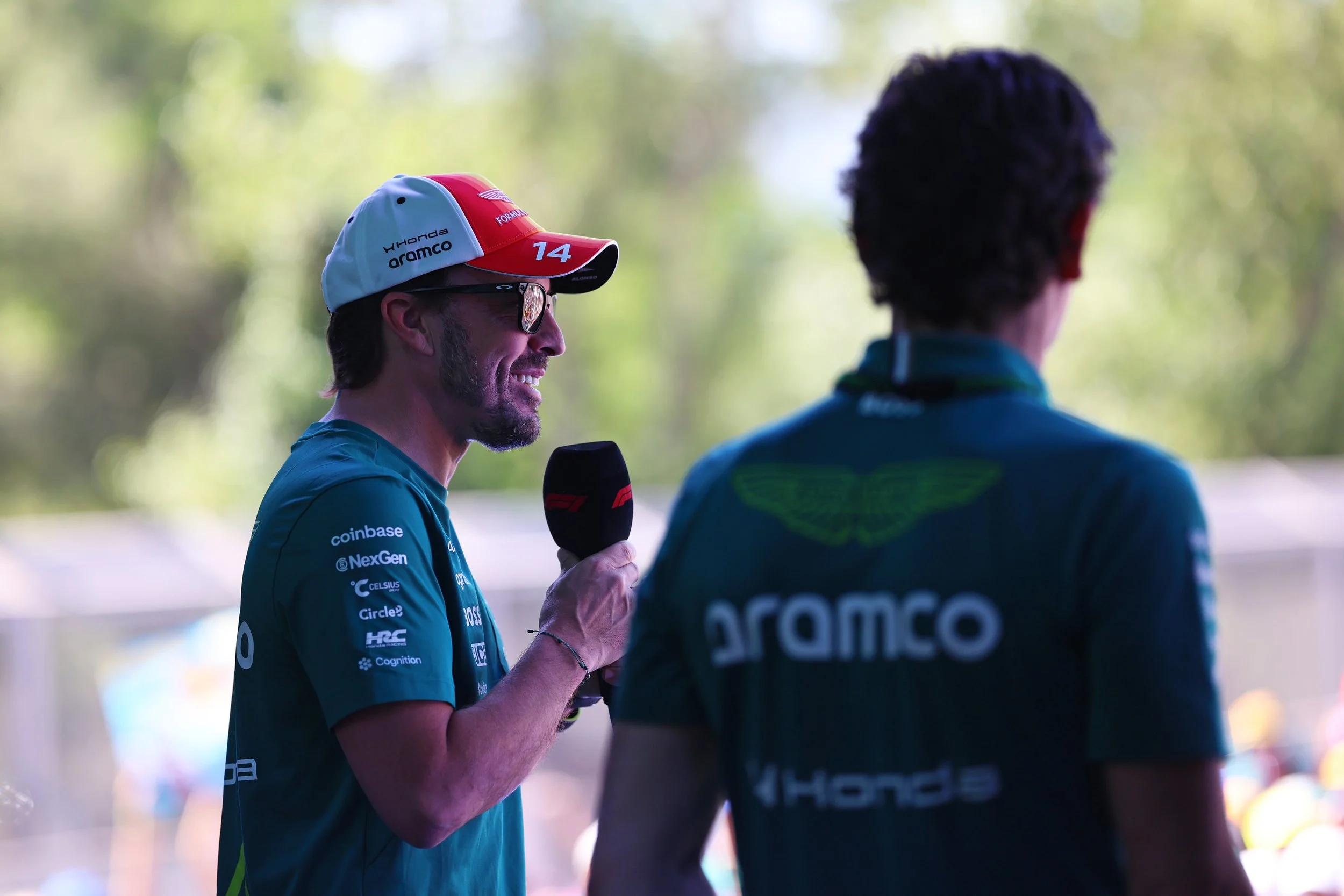 Two men in a conversation outdoors. One being Fernando Alonso, holding a microphone speaking to a crowd at the Barcelona GP 2026. The background is blurred with green trees and sunlight.