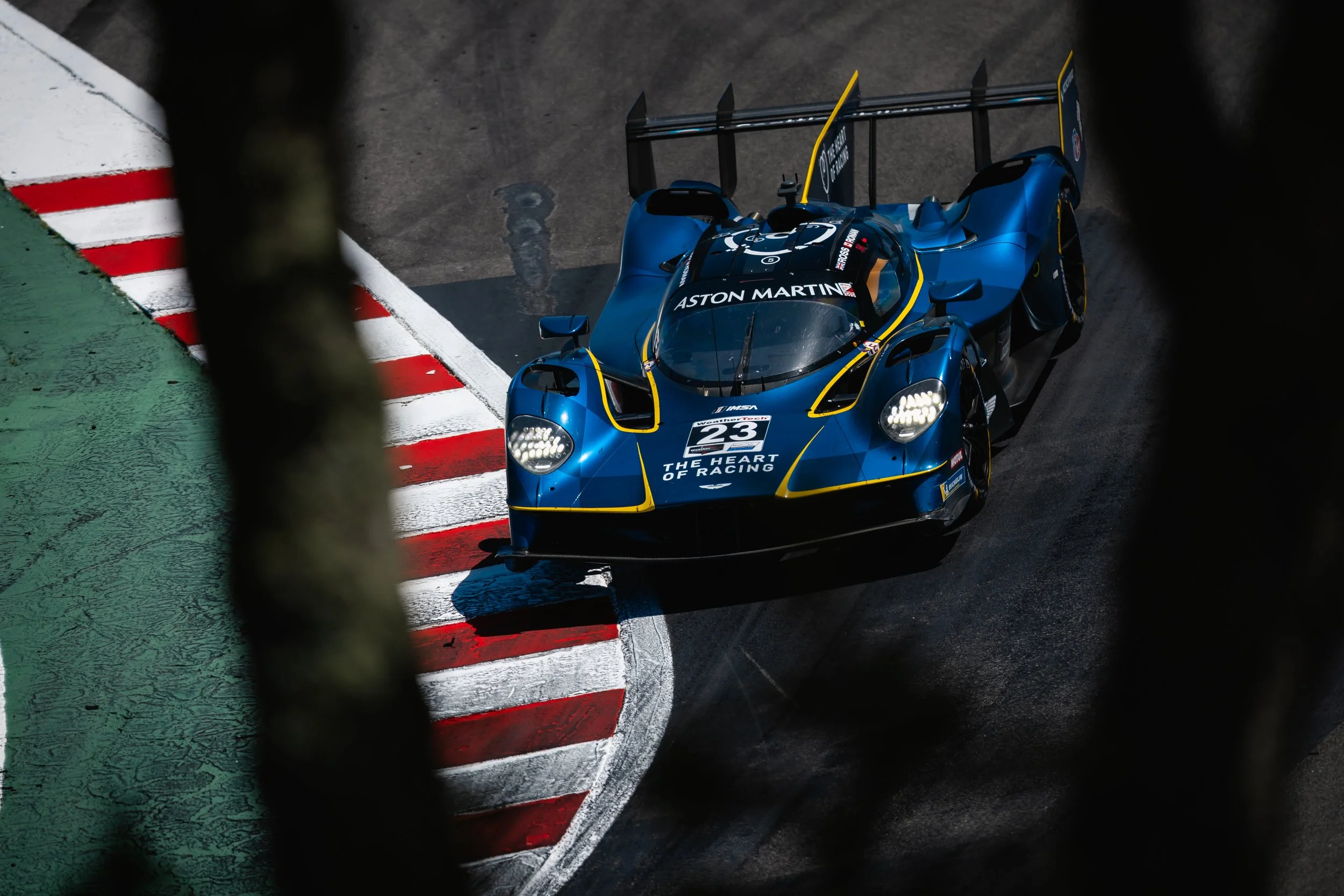 An Aston Martin Valkyrie blue race car with the number 23 and the text 'The Heart of Racing' is on a racing track, viewed through a black barrier or fence.