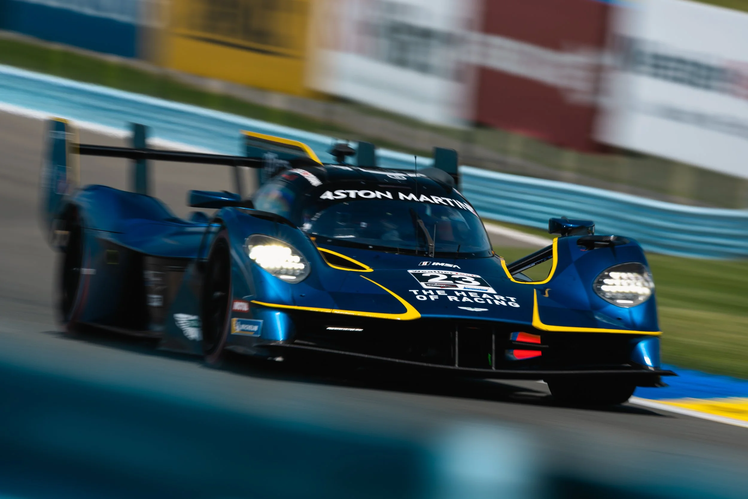 A race car with a blue and black body, yellow accents, and sponsor logos, speeding on a race track, with motion blur in the background.