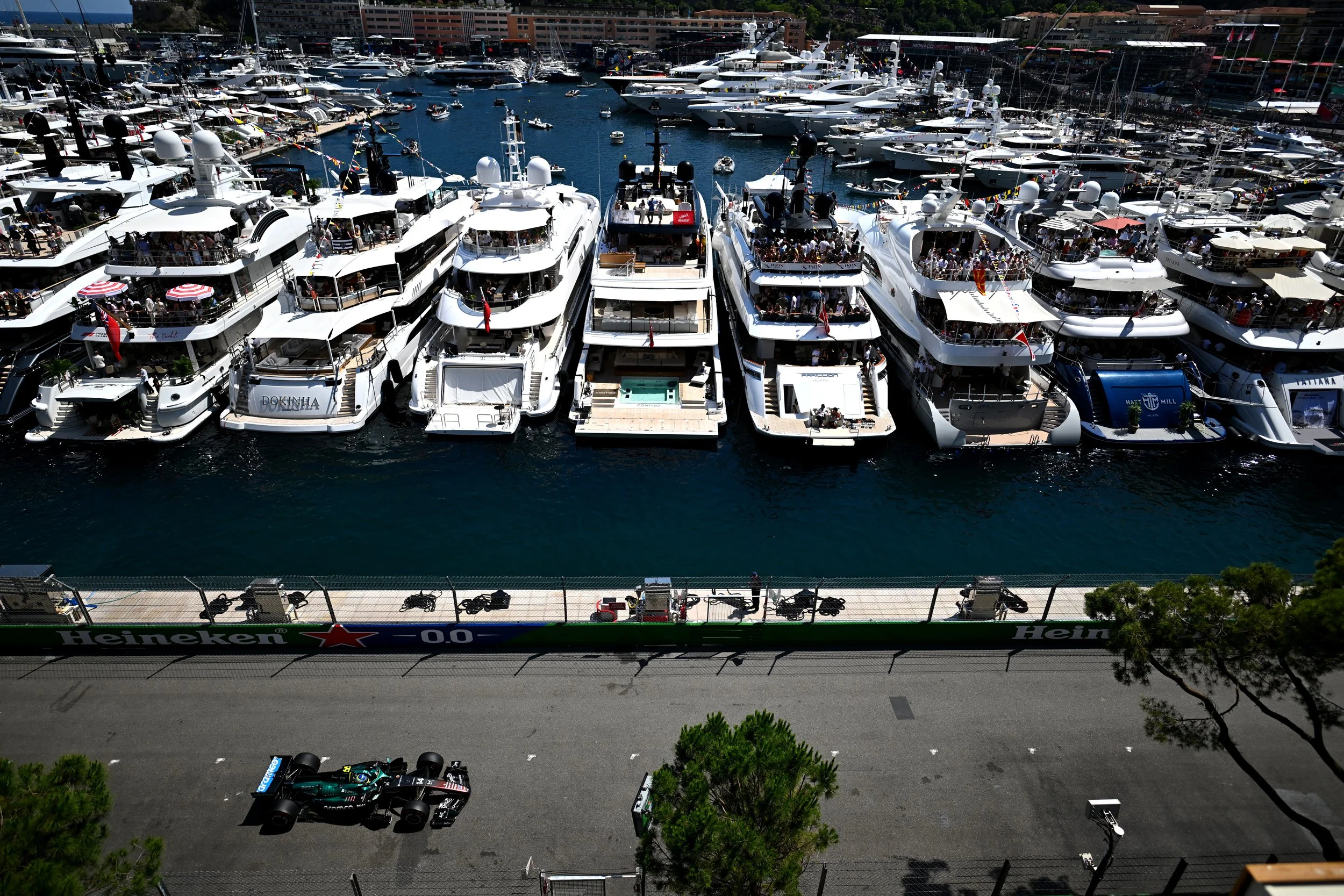 Aerial view of numerous yachts and boats docked at Monaco marina with a race track in the foreground. The marina is filled with large white yachts, some with people on board, and smaller boats scattered around. AMR26 Formula 1 Race Car is on track.