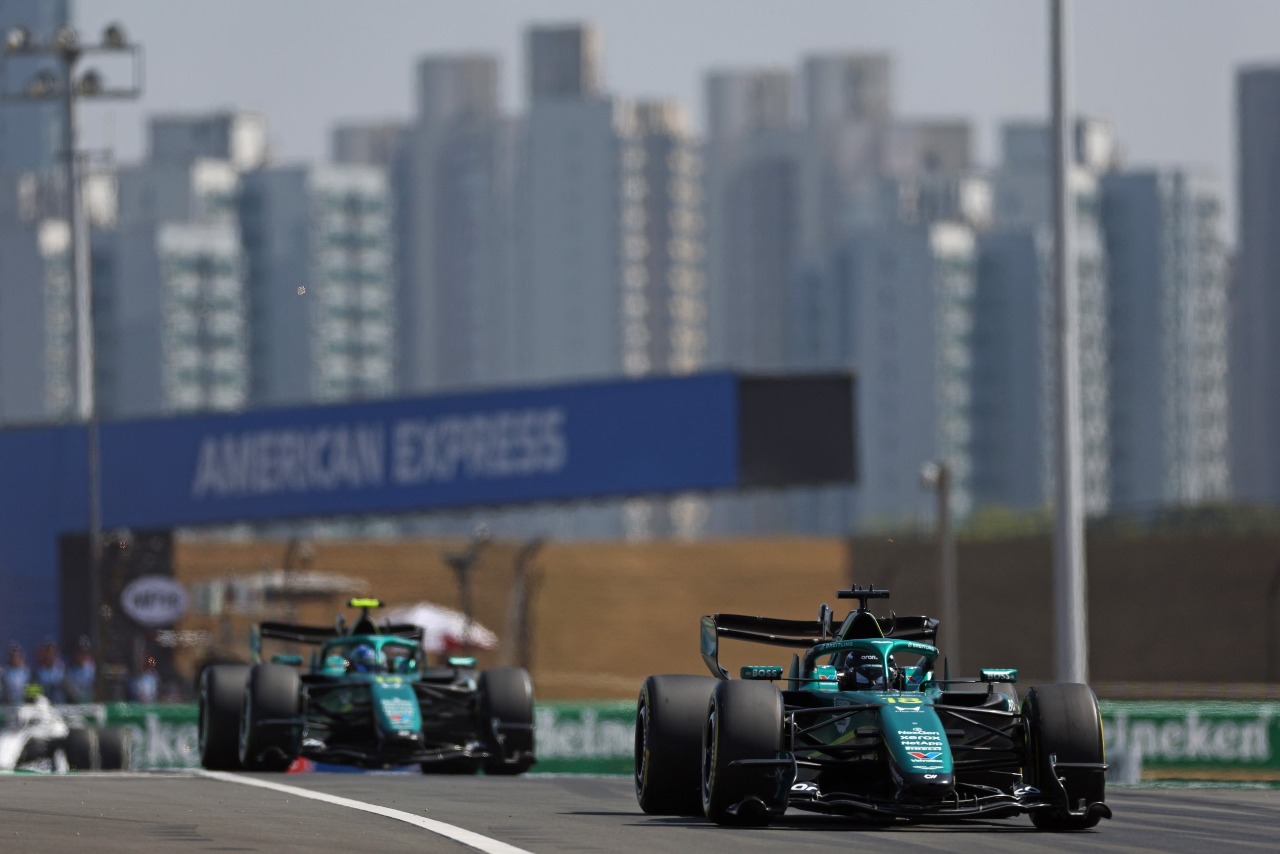 Two racing cars on a track with a blue sign reading 'AMERICAN STATES' in the background, blurred cityscape with high-rise buildings, during a professional race event.