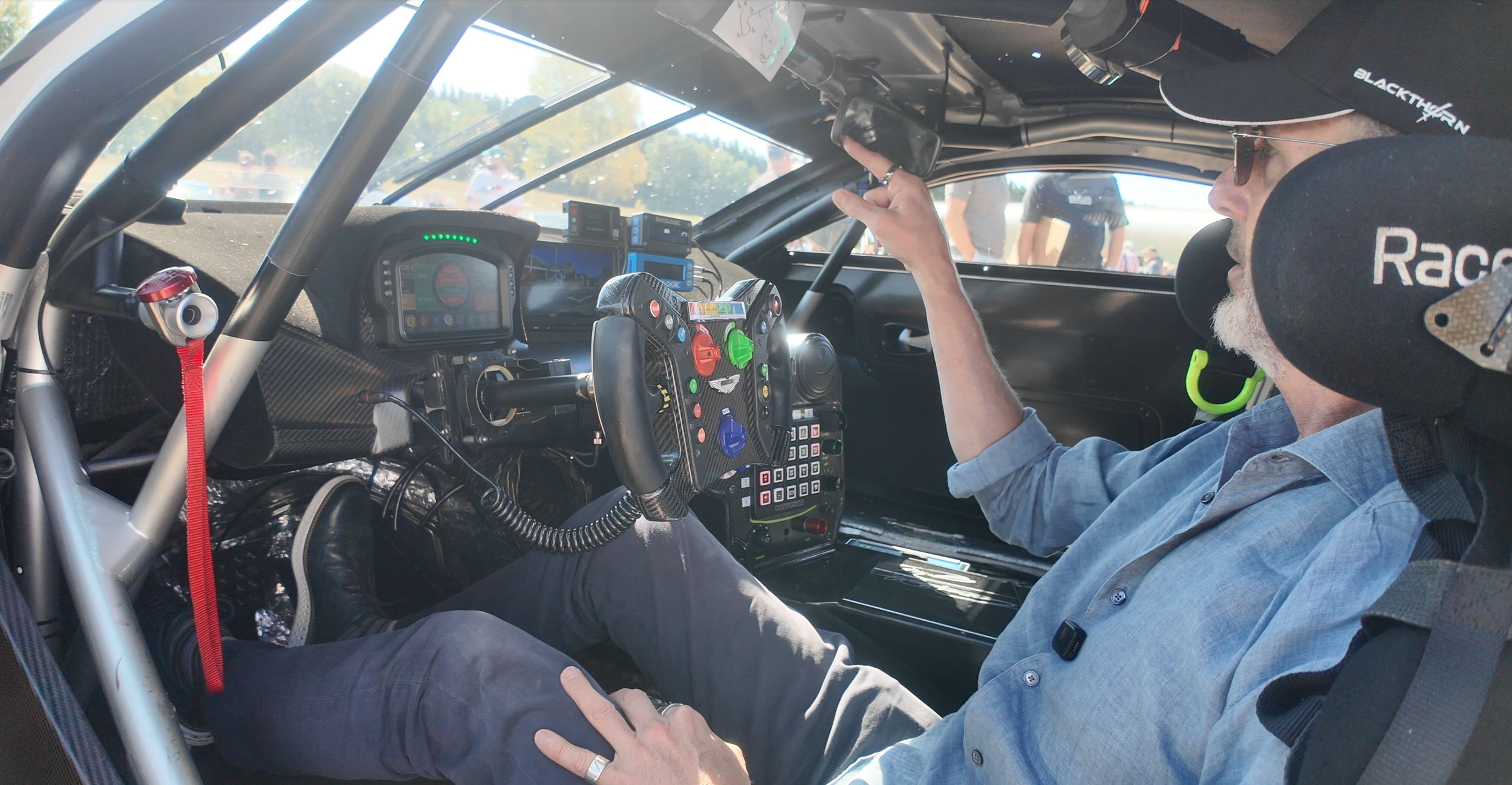 Inside a race car with a man seated in the driver seat wearing a cap, sunglasses, and a blue shirt, reaching for a monitor. The car's interior features a carbon fiber dashboard with a digital display, a racing wheel with various buttons and switches, and a roll cage. Outside, a few people are visible through the windshield.