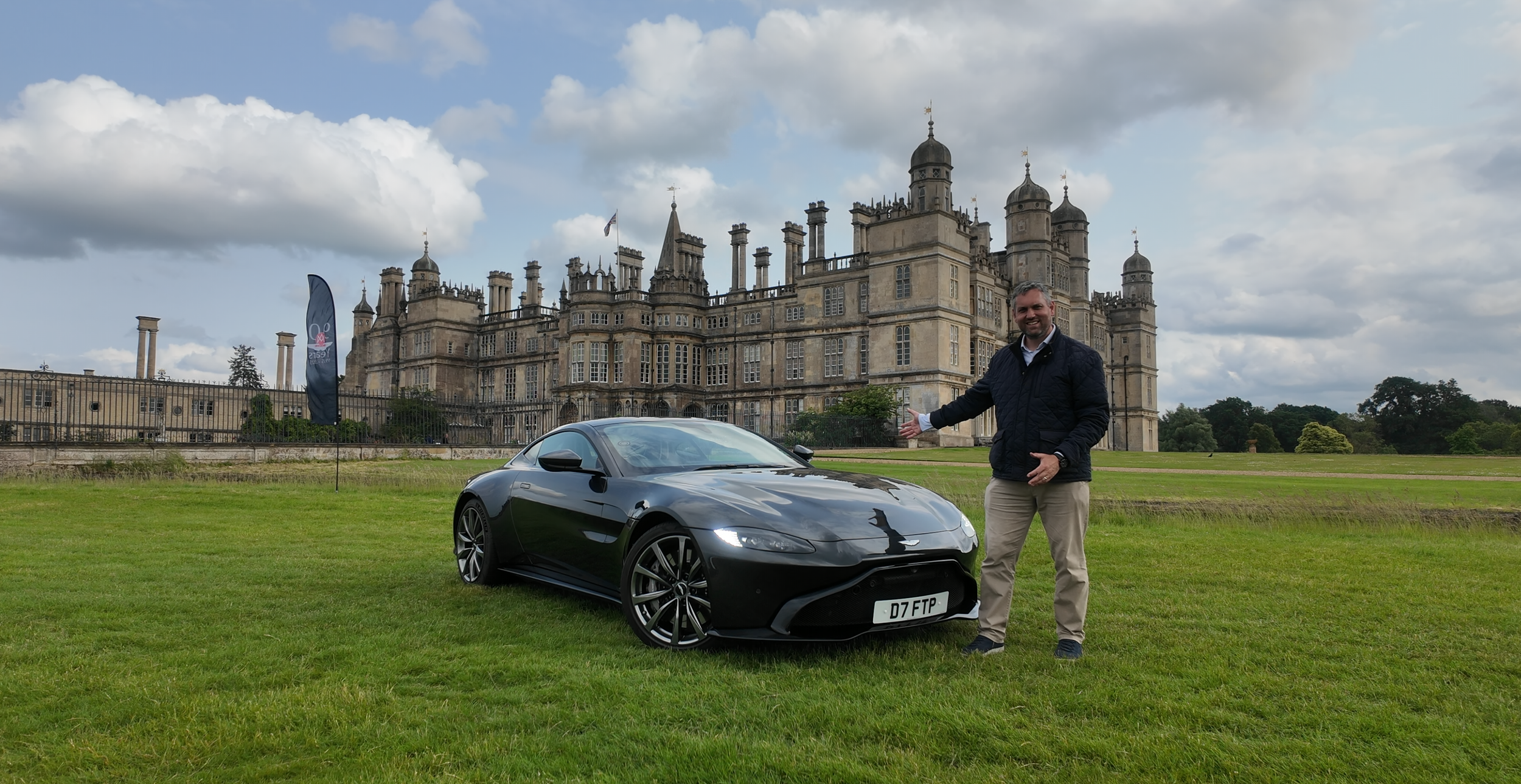A man standing next to a black sports car on a grassy field with a castle in the background and cloudy sky above.