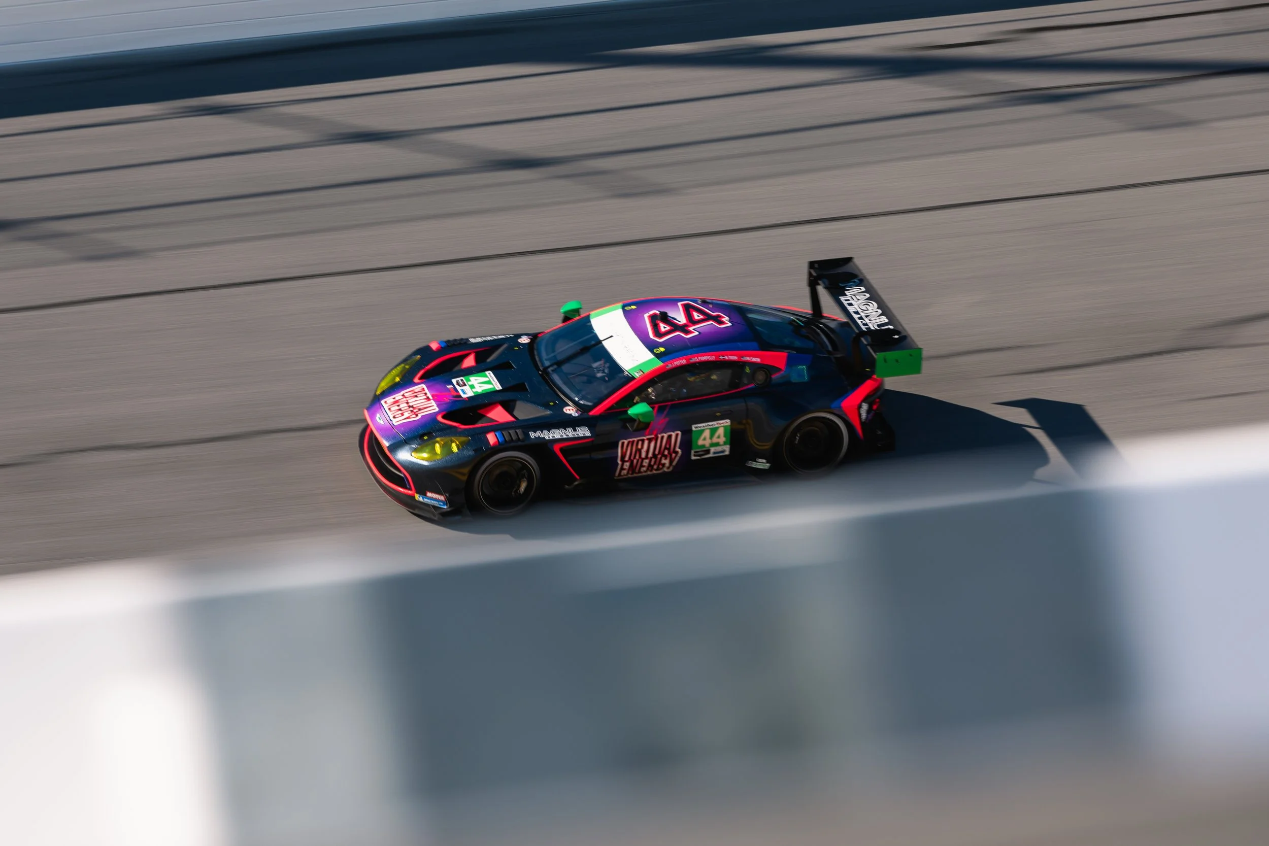 A racing car, number 44, speeding on a race track with a colorful livery and sponsor logos, including 'Virtual Energy,' as it moves rapidly, creating motion blur.