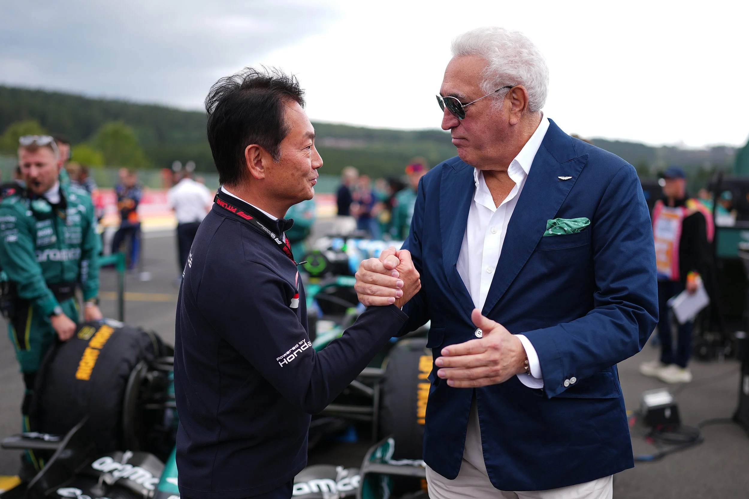 Two men shake hands on a racing track, with race cars and team members in racing suits in the background.