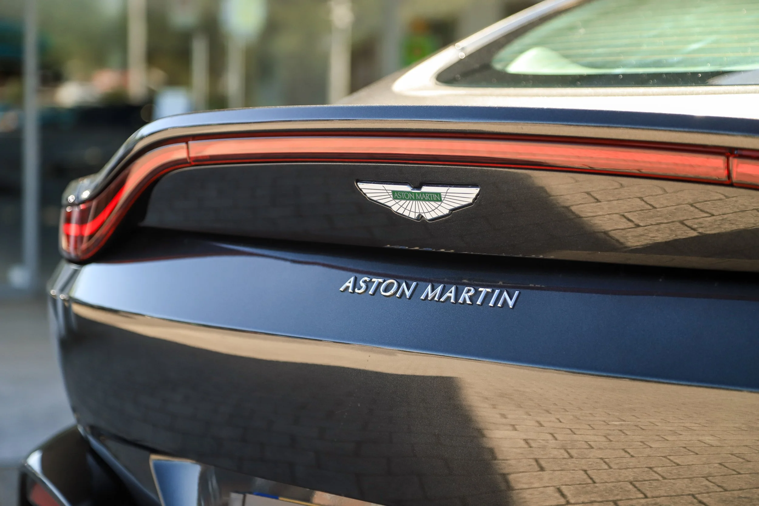 Close-up of the rear of a black Aston Martin car, showing the Aston Martin badge and logo, with part of the taillight visible.