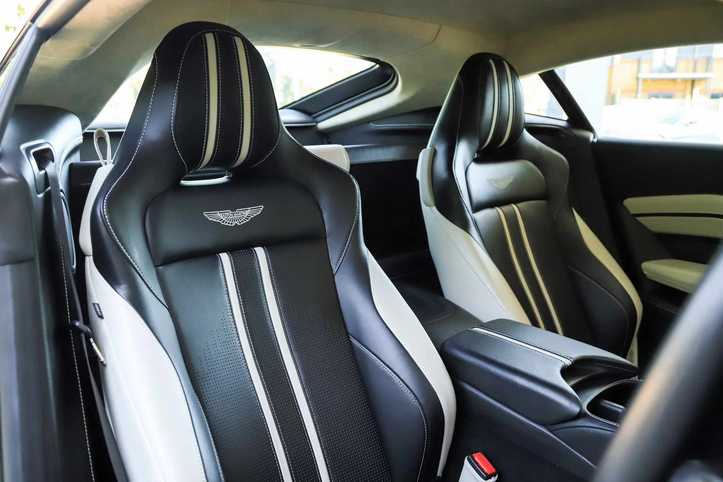 Inside the cockpit of an Aston Martin, showing two black and white racing-style seats with the Aston Martin logo, a center console, and the side window.