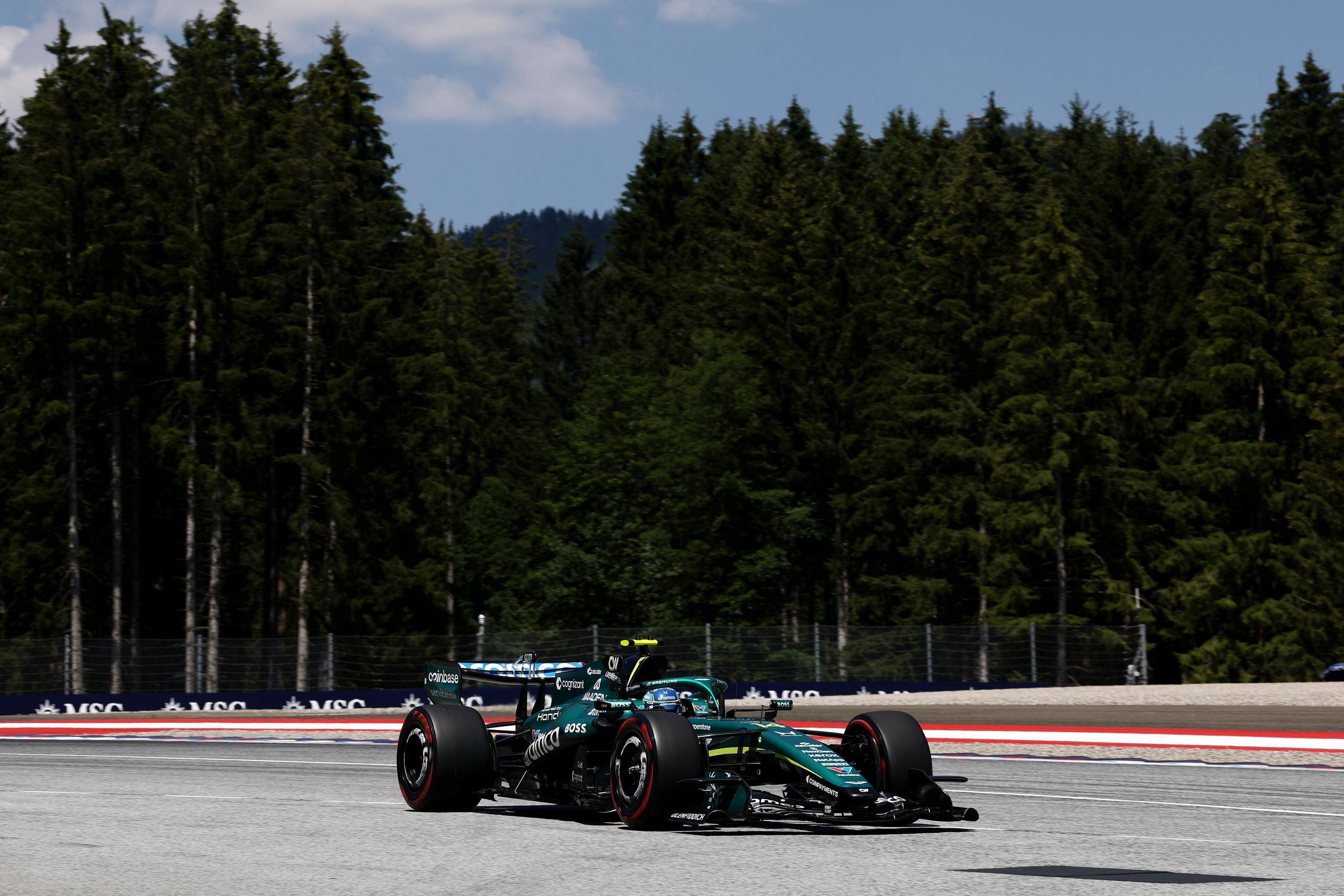 A teal and black Formula 1 race car speeding on a race track surrounded by tall green trees and a blue sky.