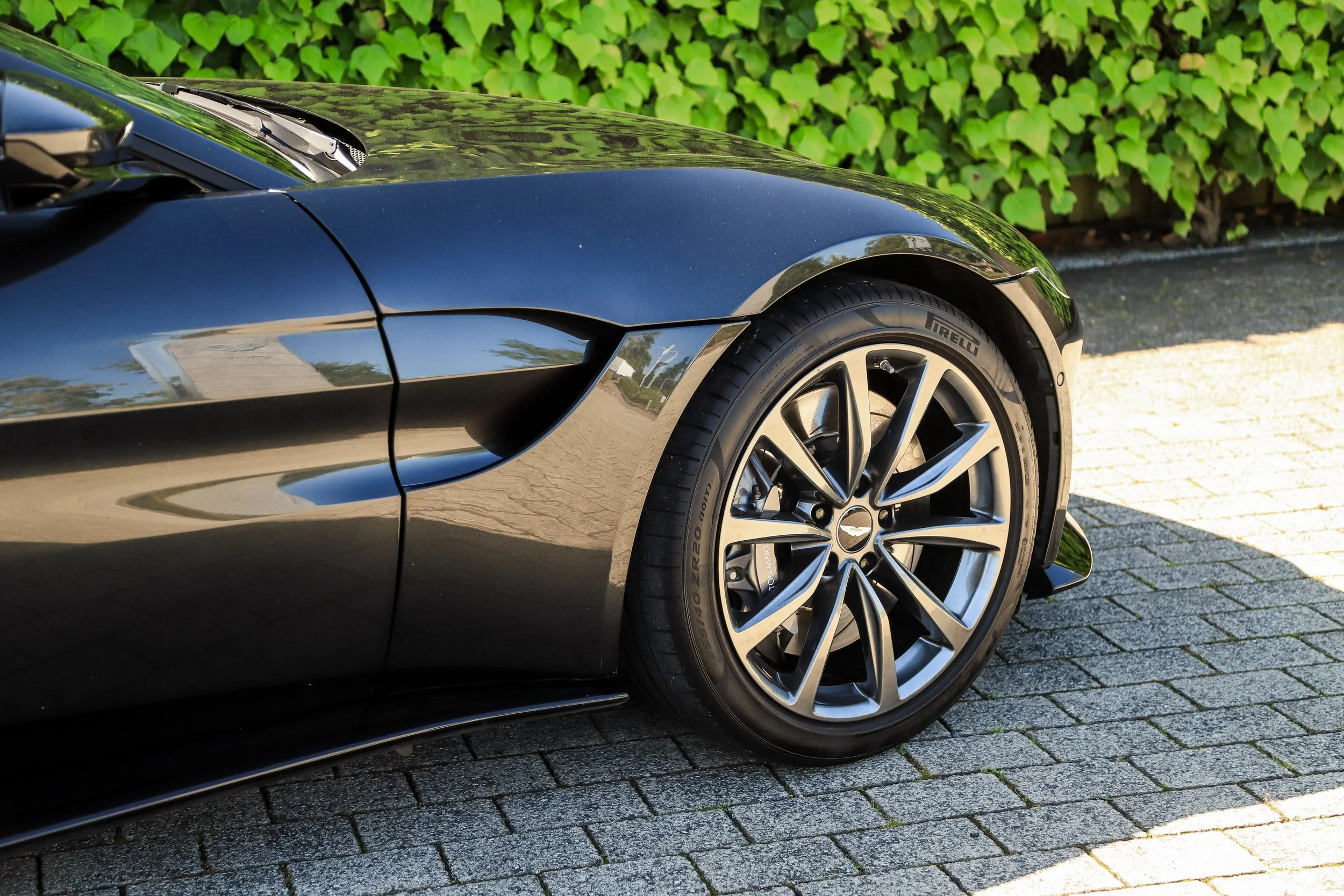 Close-up of a black sports car's front right side, showing the wheel, tire, and part of the car's body, parked on a cobblestone driveway with greenery in the background.