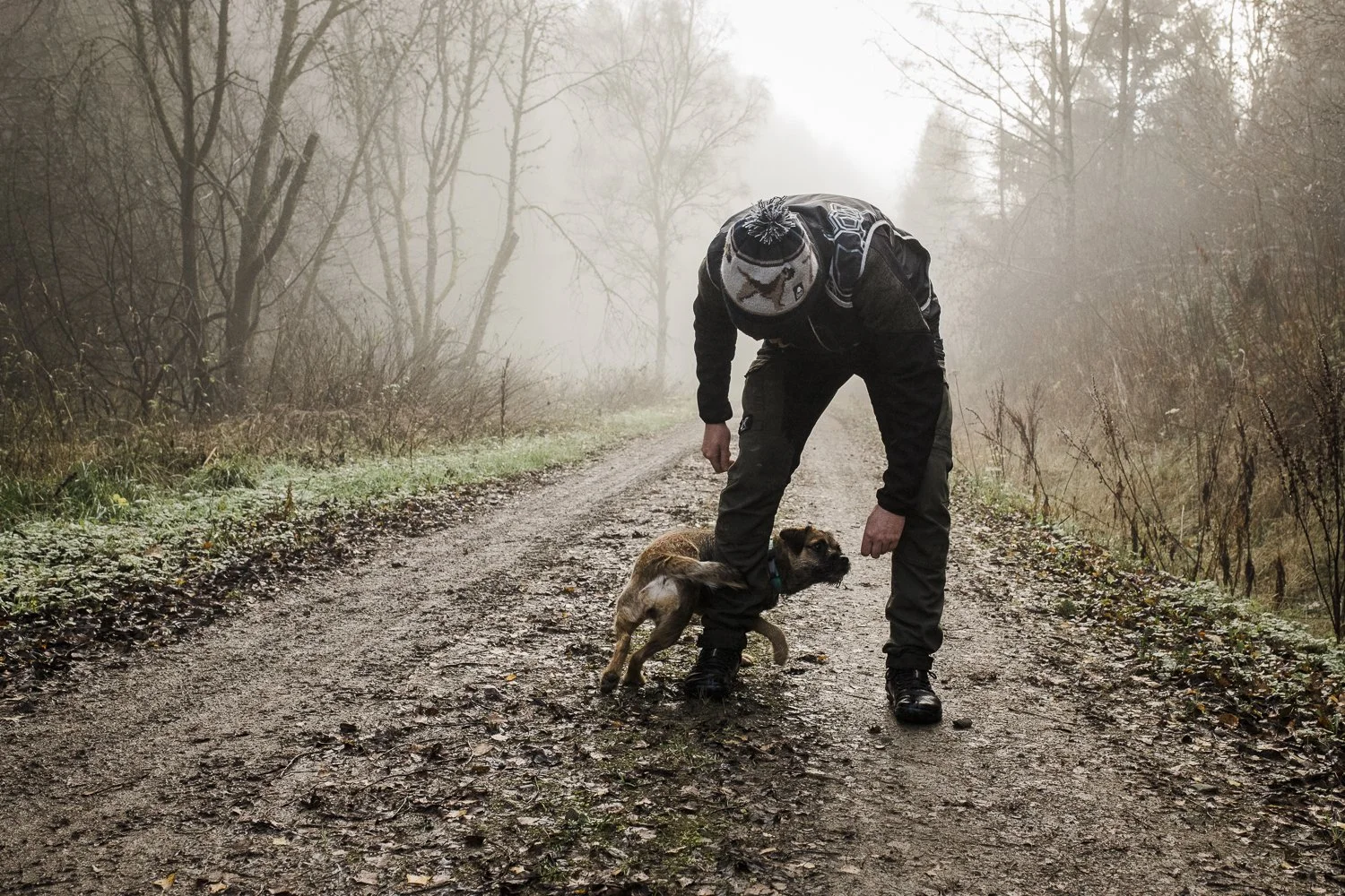 A person in outdoor gear bending down on a dirt forest trail, with a small dog between their legs, walking in a foggy, leafless wooded area.