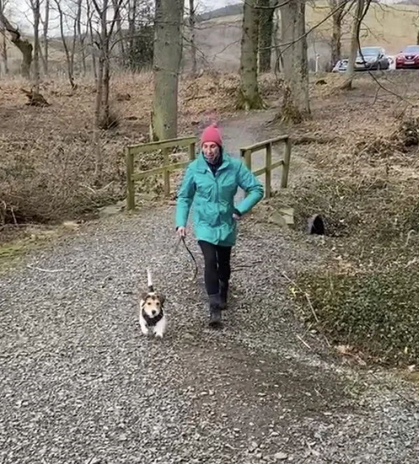 A woman in a teal jacket and pink beanie walking a small dog on a gravel path in a wooded area.